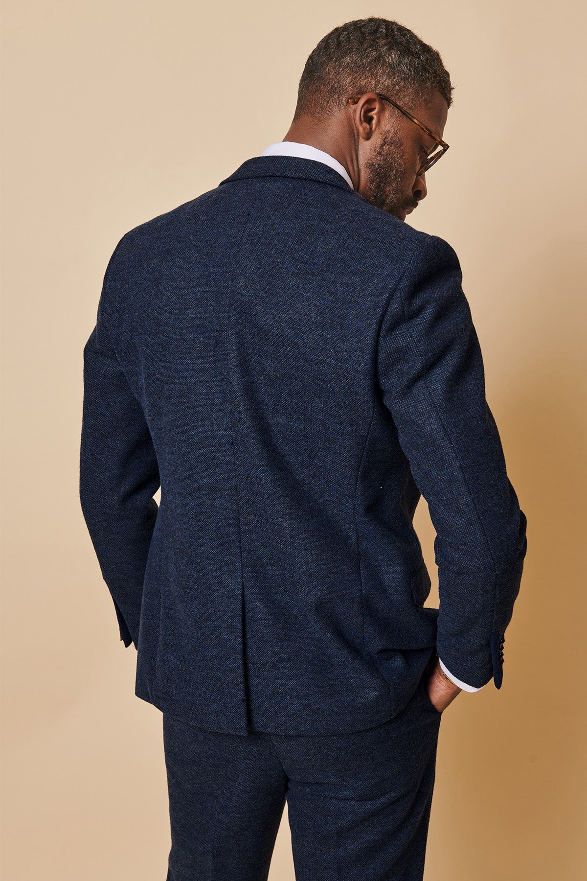 A man with short hair and glasses stands against a beige background, wearing the marcdarcy MARLOW Navy Tweed Blazer, looking over his shoulder with one hand in his pocket.