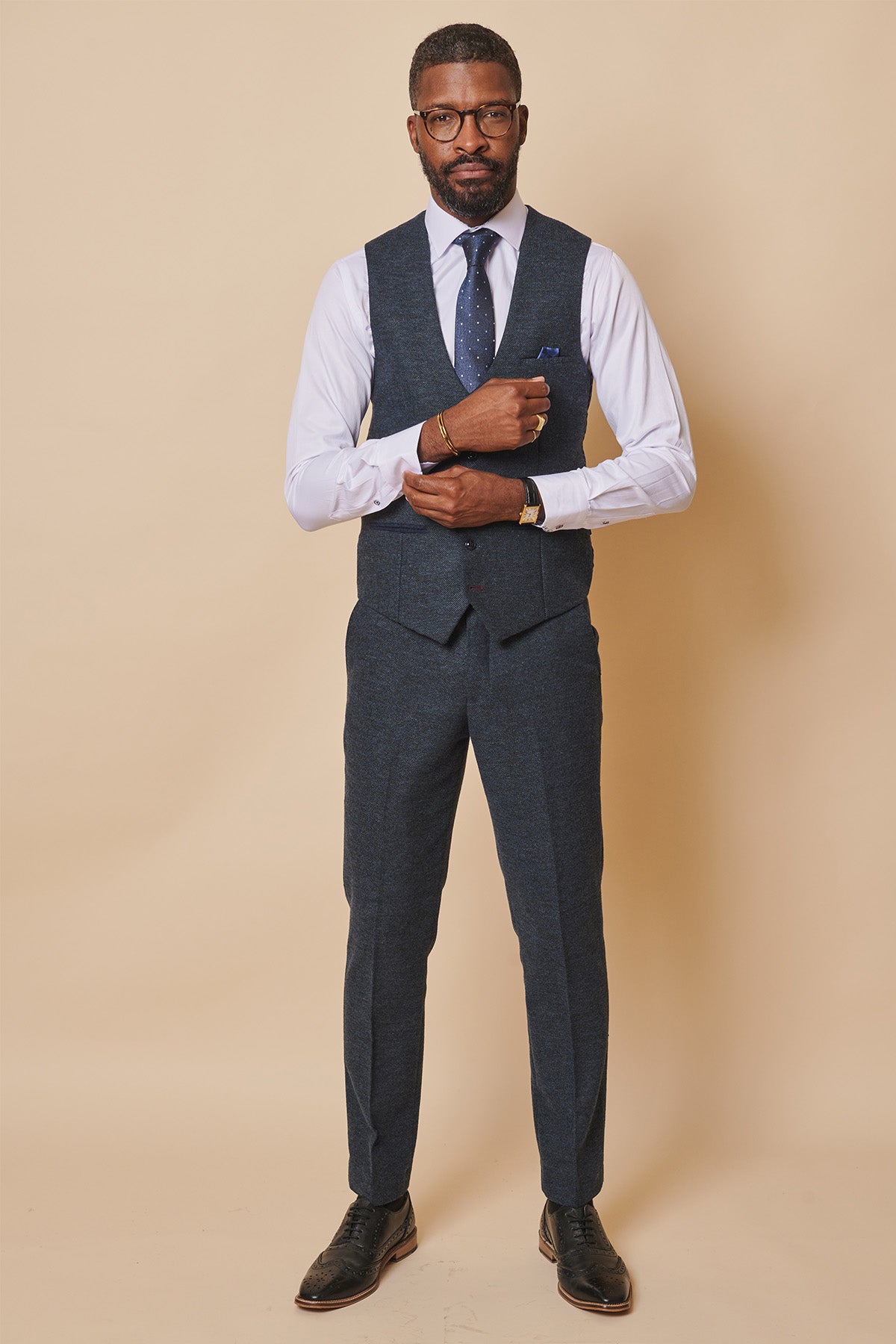 A man poses against a beige background wearing the marcdarcy MARLOW Blue Tweed Single Breasted Waistcoat, paired with matching pants, a white shirt, blue dotted tie, and black shoes. He adjusts his left cuff while looking at the camera.