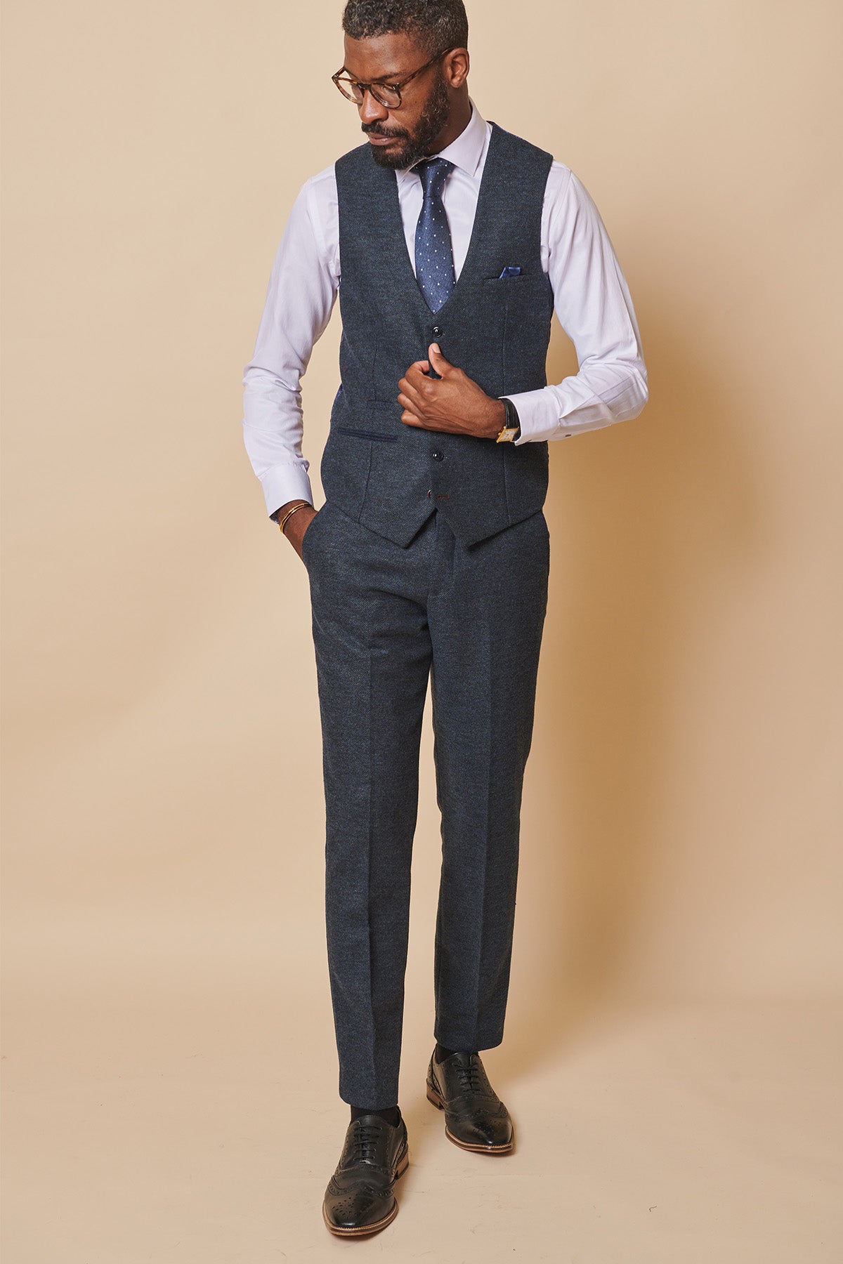 A Black man in a three-piece suit and glasses looks down, one hand in his pocket while touching his marcdarcy MARLOW blue tweed single breasted waistcoat, made from poly wool.