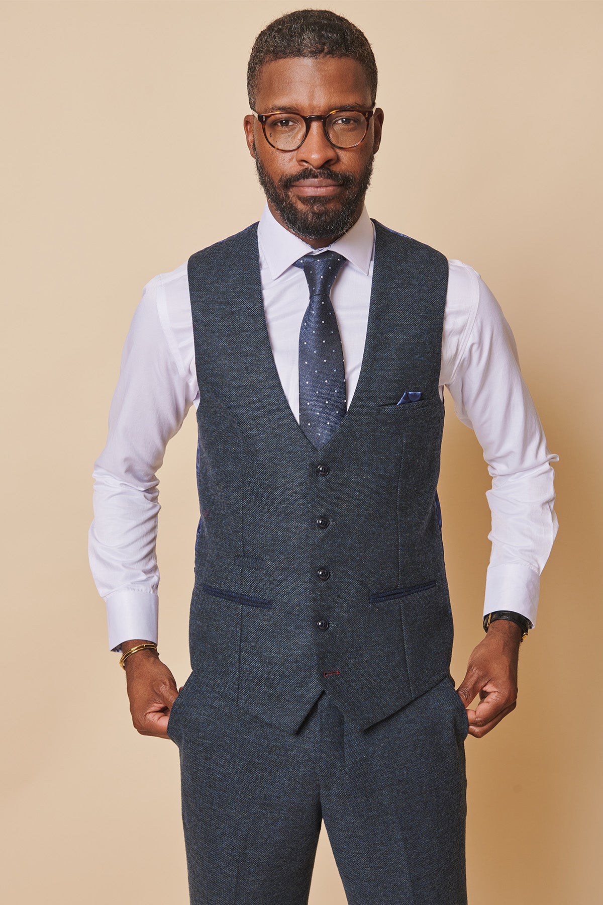 A man with short hair and a beard wears glasses, a white dress shirt, the marcdarcy MARLOW Blue Tweed Single Breasted Waistcoat, matching trousers, and a navy polka dot tie, standing against a beige background with hands in his pockets.