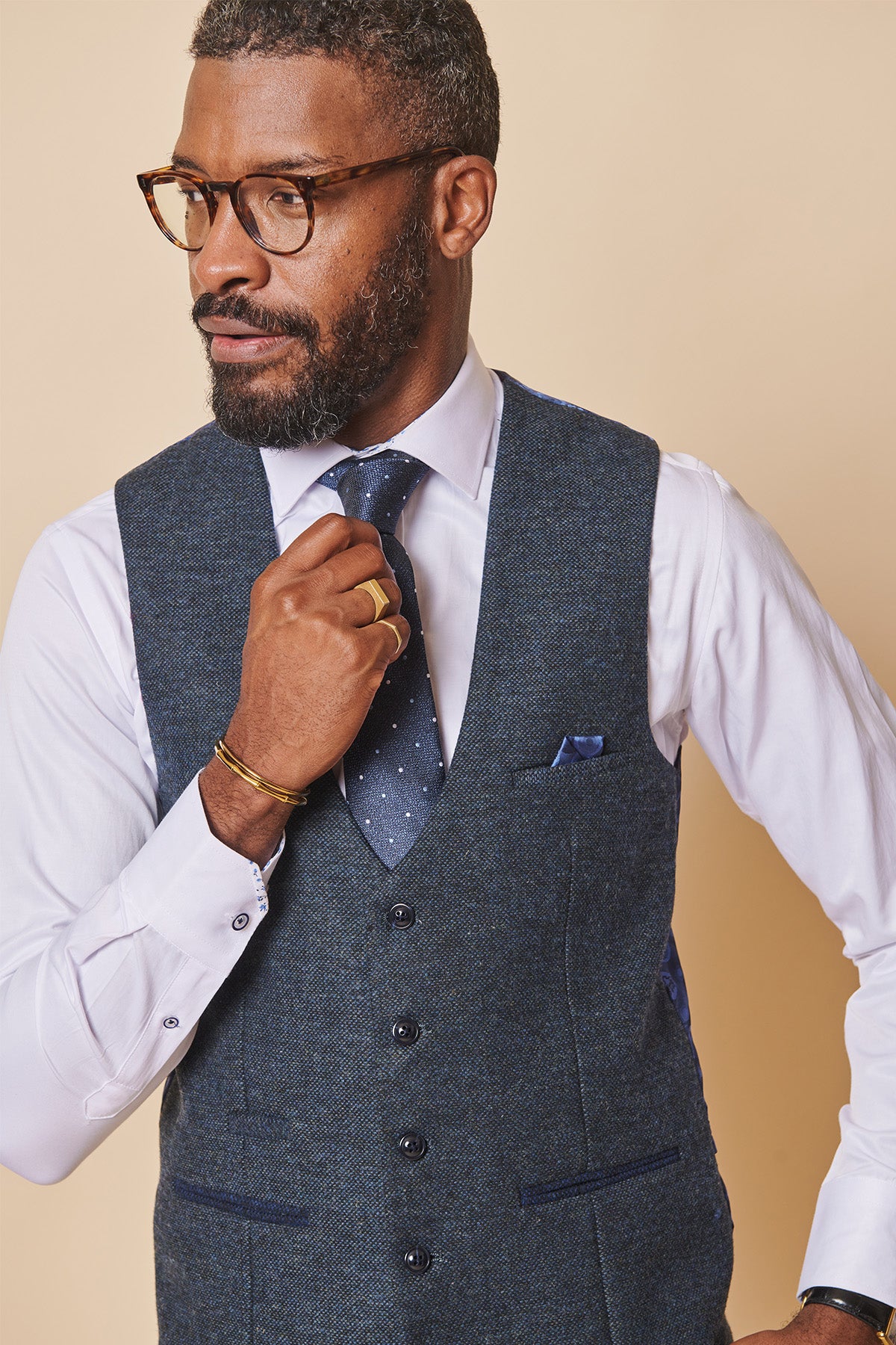 A man with short curly hair, a beard, and glasses adjusts his blue tie. He wears a white shirt and the marcdarcy MARLOW Blue Tweed Single Breasted Waistcoat (slim fit) against a plain light beige background.