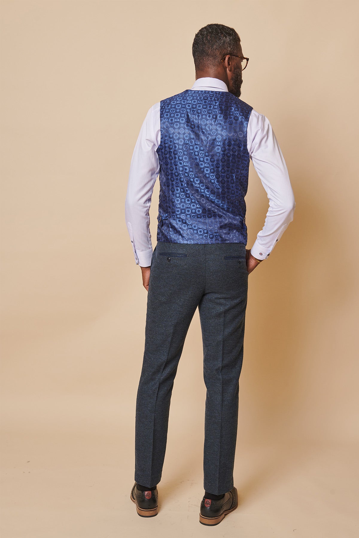 A man faces away, wearing a white shirt, dark gray trousers, brown shoes, and the marcdarcy MARLOW Blue Tweed Single Breasted Waistcoat in slim fit poly wool tweed, set against a beige background.