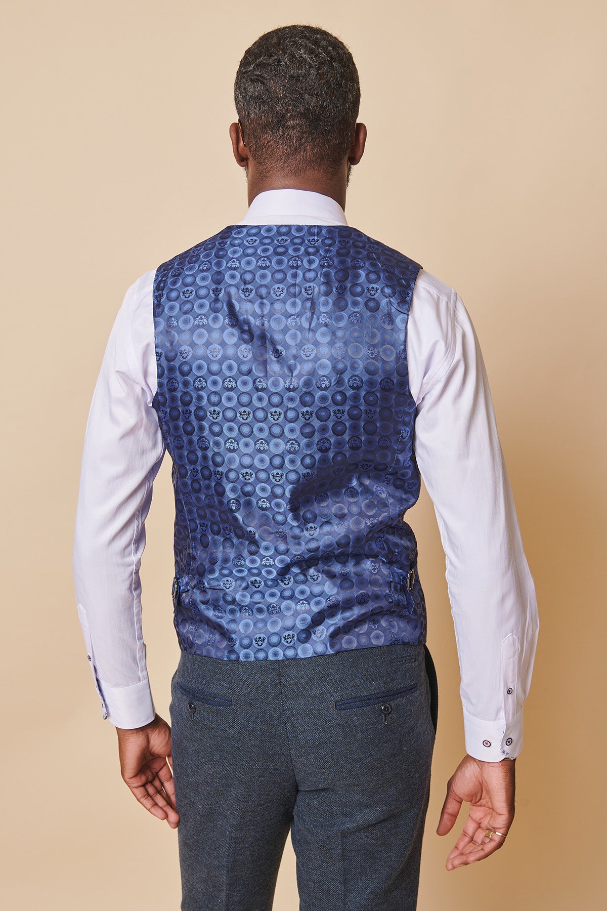 A man is seen from behind wearing a white shirt, dark grey trousers, and the marcdarcy MARLOW Blue Tweed Single Breasted Waistcoat in a slim fit with a circular pattern, standing against a beige background.