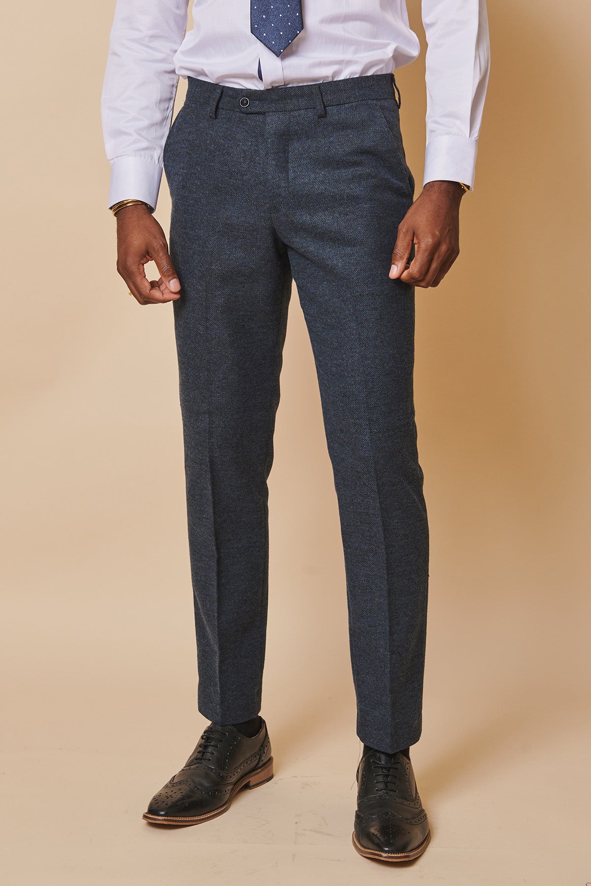 A person wears marcdarcy's MARLOW Blue Tweed Trousers with a white dress shirt, blue patterned tie, and black dress shoes against a beige background. The image is cropped at the shoulders.