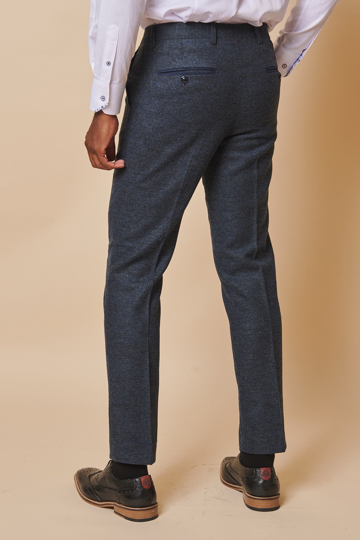 A person wearing marcdarcy MARLOW Blue Tweed Trousers, a white shirt, black shoes, and black socks stands facing away from the camera against a beige background.