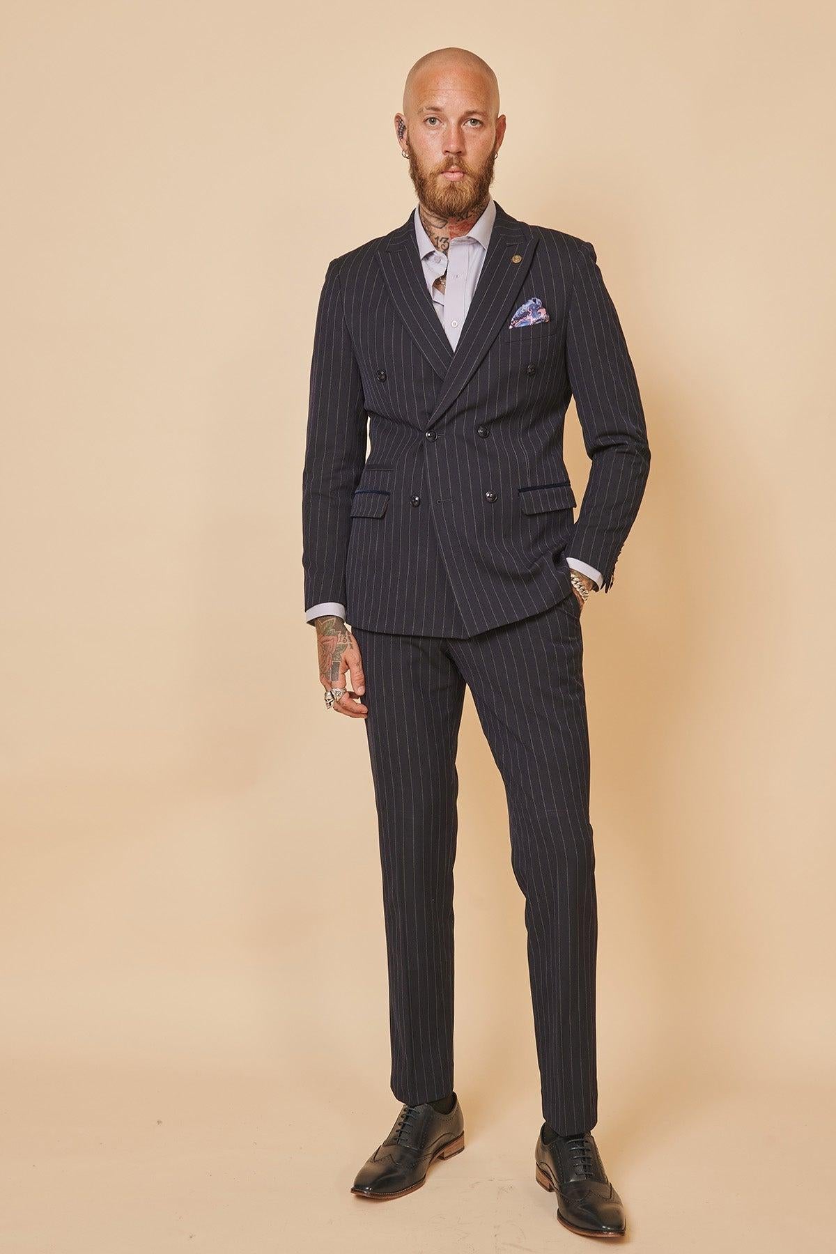 A bald, bearded man with tattoos stands against a plain background wearing the Marc Darcy Cardiff City F.C. Collection - ROCCO Navy Double Breasted Suit as worn by Ryan Wintle, complete with a pocket square.