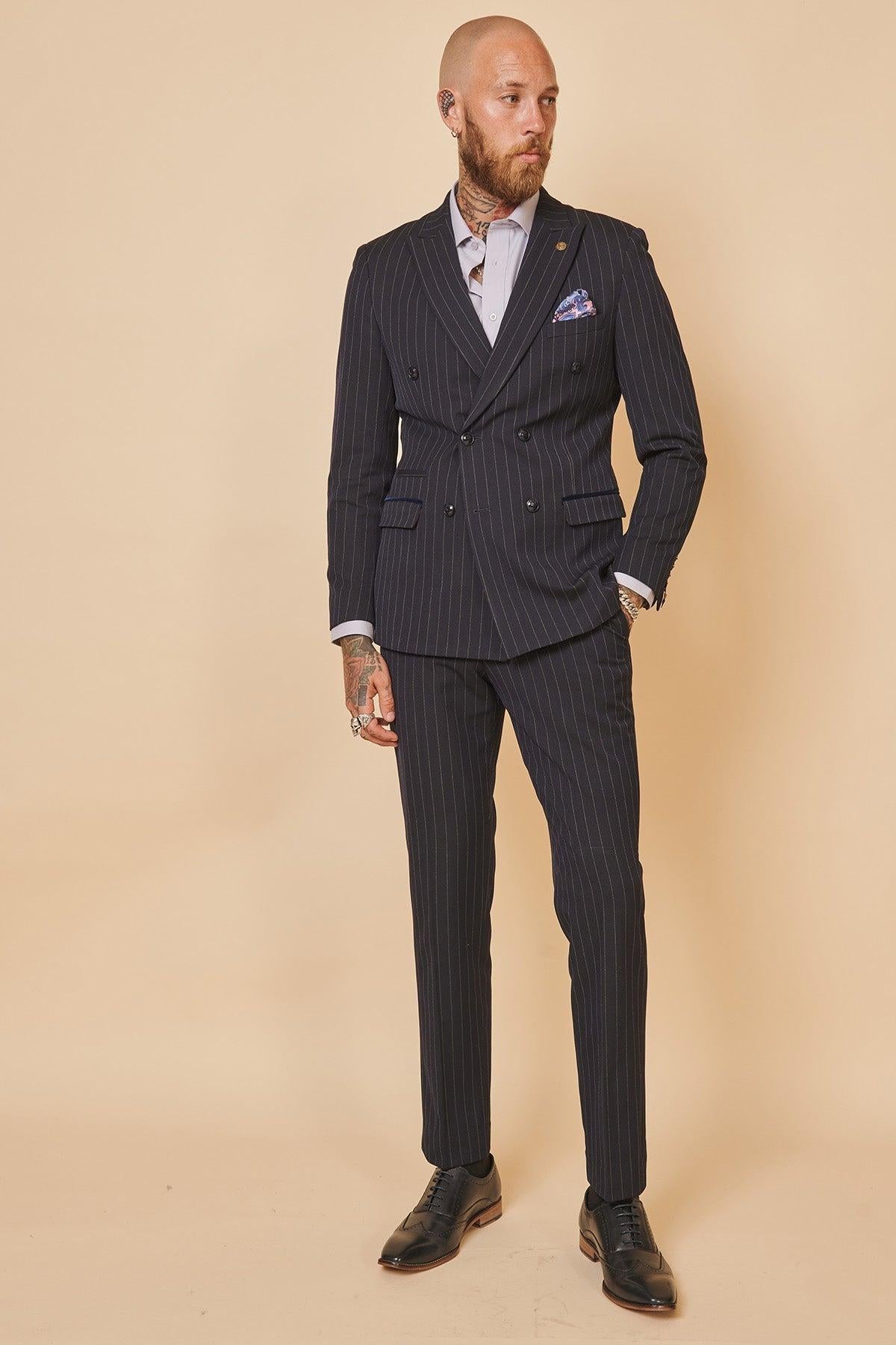 A bald, tattooed man wears the Marc Darcy Cardiff City F.C. Collection – ROCCO Navy Double Breasted Suit as worn by Ryan Wintle, paired with a light dress shirt and black shoes, standing against a beige backdrop.