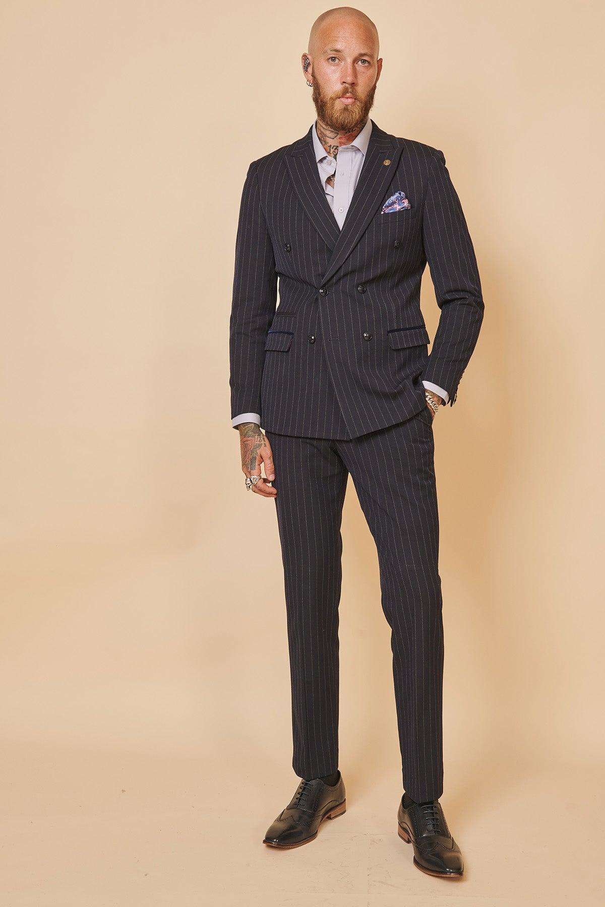 Bald man with a beard stands confidently against a beige background wearing The Everton Collection | Jordan Pickford In ROCCO Navy Pinstripe Double Breasted Two Piece Suit by Marc Darcy, paired with a white shirt, black dress shoes, hand tattoos, and visible earrings.