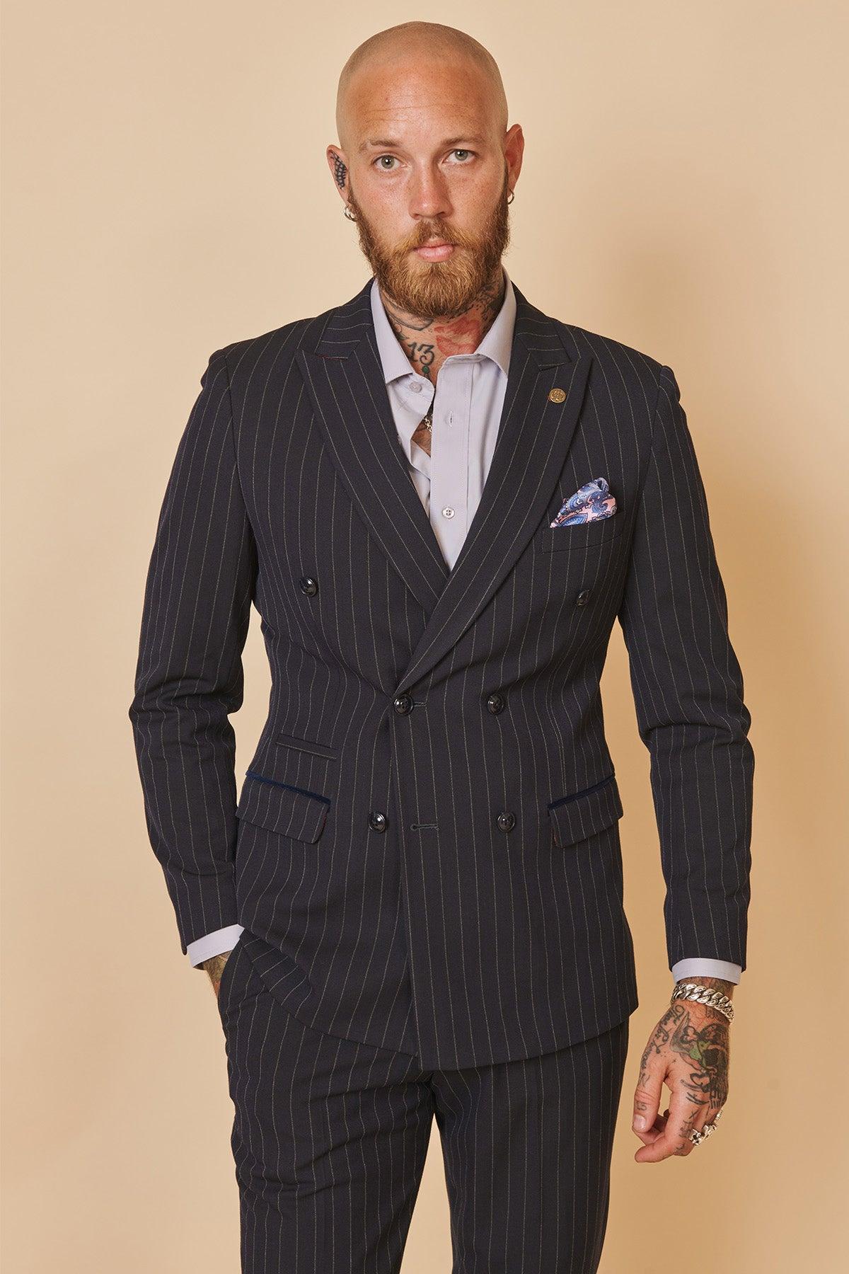 Coronation Street’s Mason Radcliffe (Luca Toolan) wears a Marc Darcy ROCCO Navy Pinstripe Double Breasted Two Piece Suit, light shirt, and pocket square. He stands against a beige background, looking at the camera with a neutral expression.