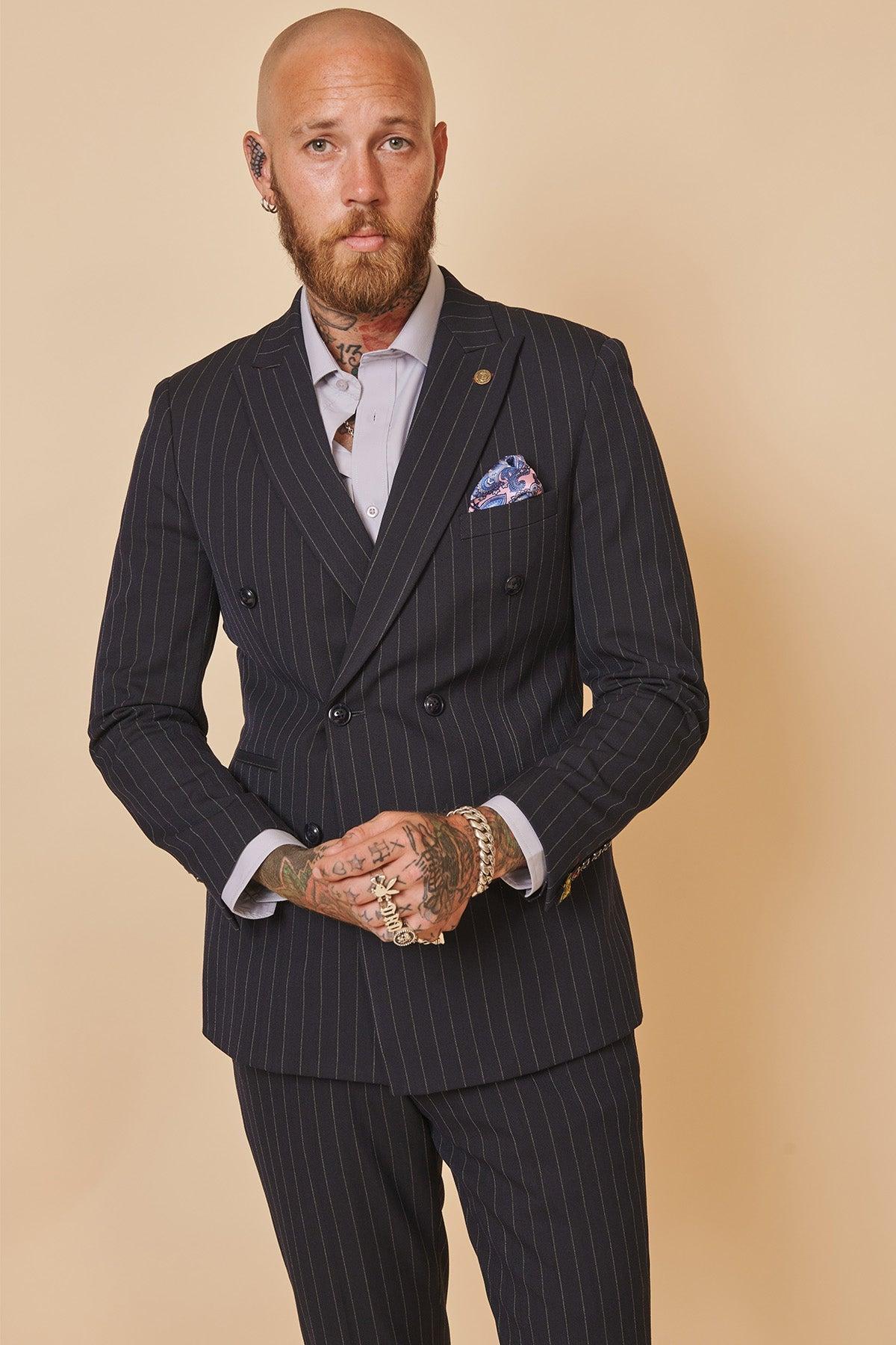 Coronation Street’s Mason Radcliffe (Luca Toolan) wears the ROCCO navy pinstripe double breasted two piece suit by Marc Darcy, styled with a white shirt and patterned pocket square against a beige background.