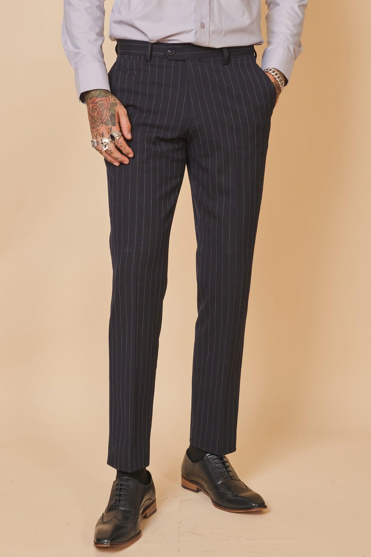 Man wears the Marc Darcy "Everton Collection | Jordan Pickford in ROCCO Navy Pinstripe Double Breasted Two Piece Suit," styled with a light shirt, black socks, oxford shoes, rings, and bracelet—showcasing modern slim fit menswear. Tattoos on his left hand add edge.