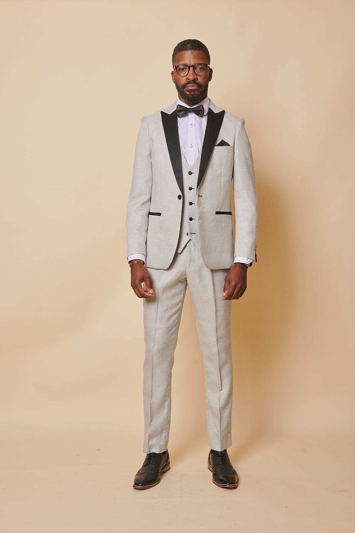 A man wearing the marcdarcy SPENCER Stone Tux Lapel Three Piece Suit stands against a beige background with a neutral expression, styled with a white shirt, black bow tie, pocket square, glasses, and black dress shoes.
