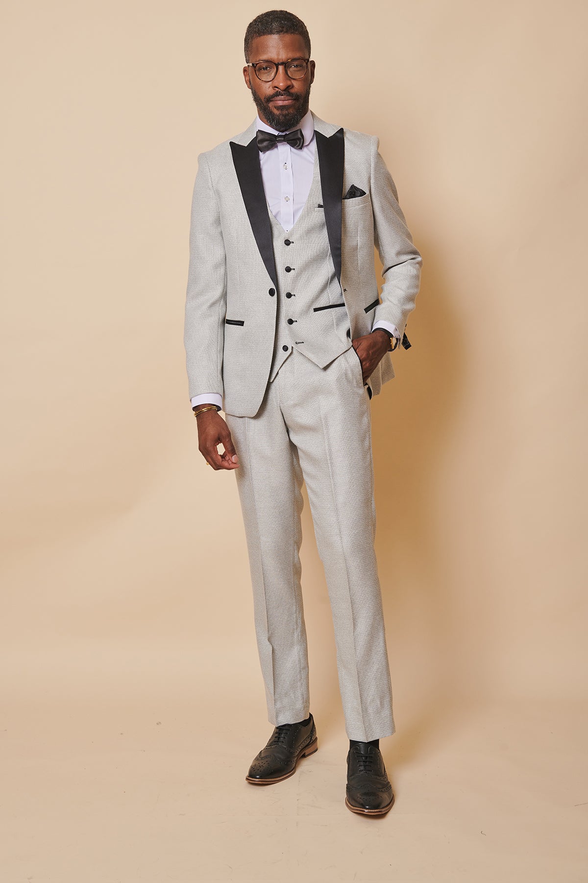 A bearded man with glasses stands against a beige background in the marcdarcy SPENCER Stone Tux Lapel Three Piece Suit, styled with a white shirt, black dress shoes, and a pocket square—ideal for formal occasions. One hand is in his pocket.