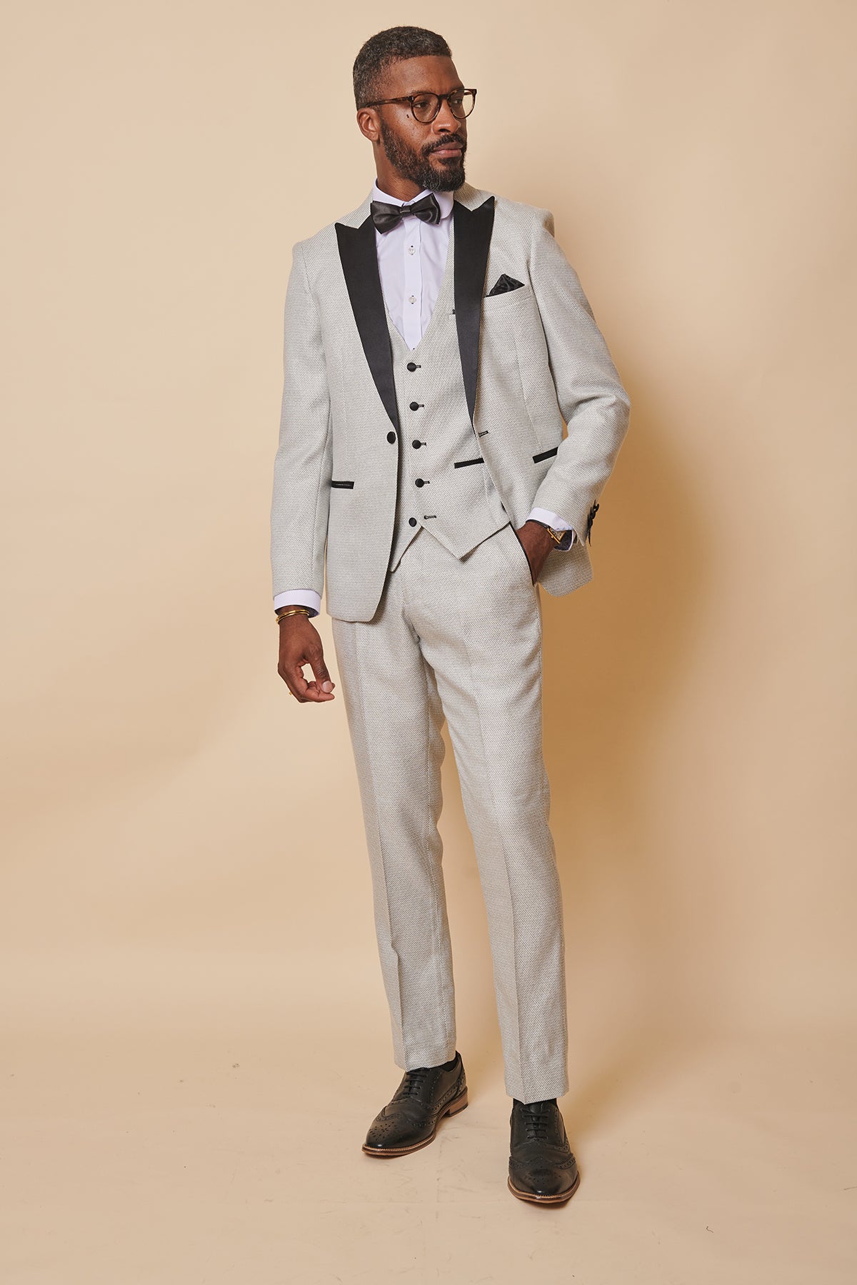 A man wears the marcdarcy SPENCER Stone Tux Lapel Three Piece Suit with black lapels, a black bow tie, white shirt, and black dress shoes, standing confidently against a beige background—ideal for formal occasions.
