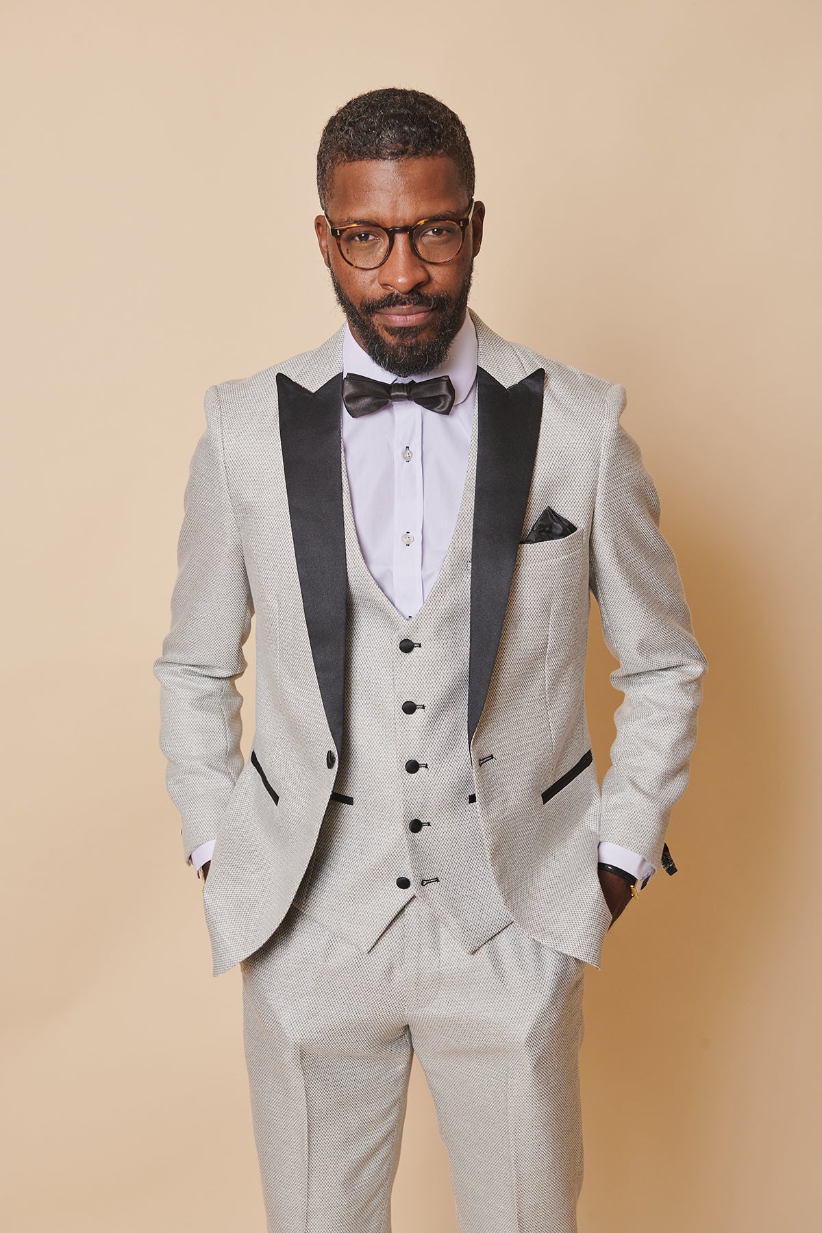 A man wears the marcdarcy SPENCER - Stone Tux Lapel Blazer with black lapels, a white shirt, and a black bow tie against a beige background—an ideal look for black tie events.