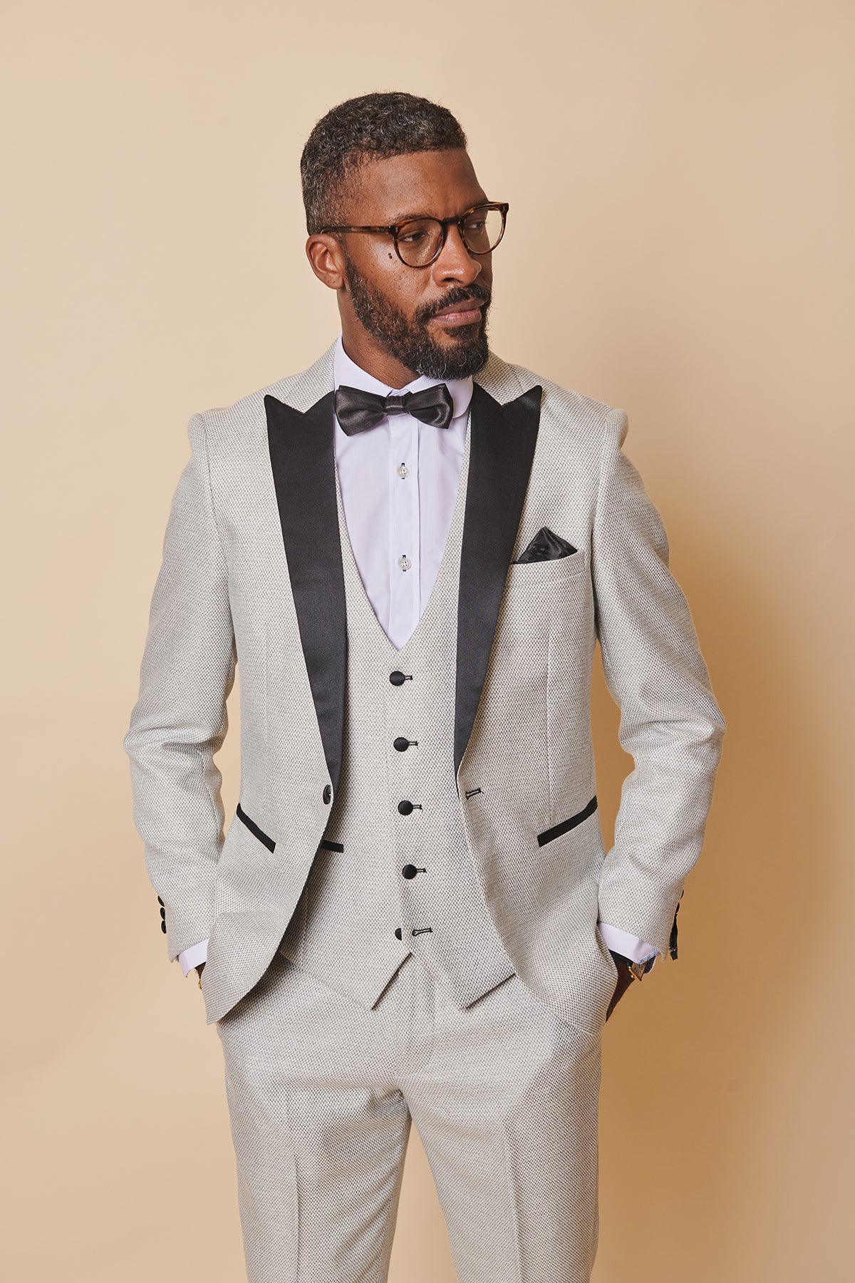 A bearded, short-haired man in glasses wears the marcdarcy SPENCER - Stone Tux Lapel Blazer with black lapels, a white shirt, black bow tie, and vest, standing hands-in-pockets against a beige background—ideal for black tie events.