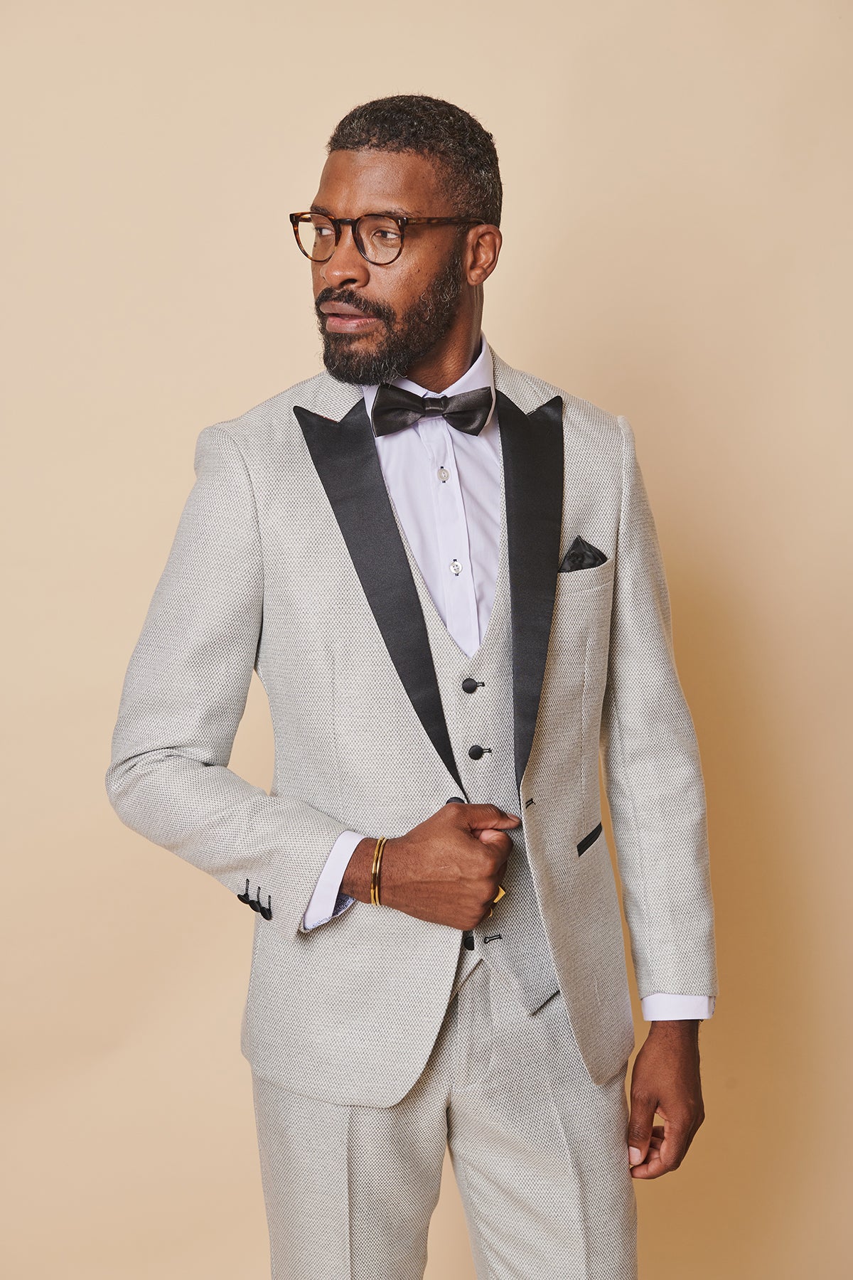 A bearded man in glasses wears the marcdarcy SPENCER Stone Tux Lapel Blazer with a white shirt, black bow tie, and pocket square, standing against a beige background—an ideal look for black tie events.