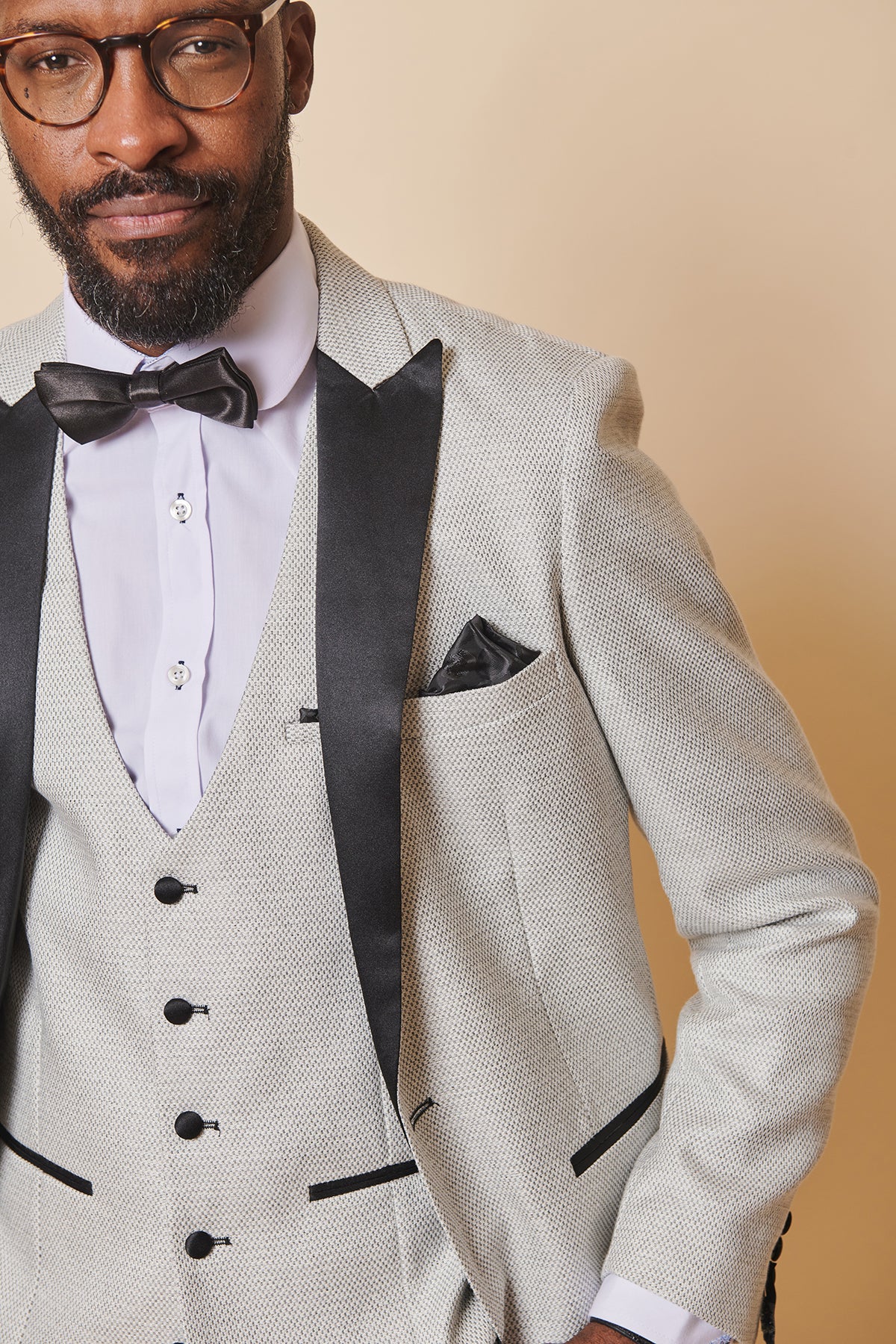 A man wears the marcdarcy SPENCER Stone Tux Lapel Blazer with black lapels, a black bow tie, and a white shirt. He has glasses, a neat beard, and stands against a beige background—ideal for black tie events.