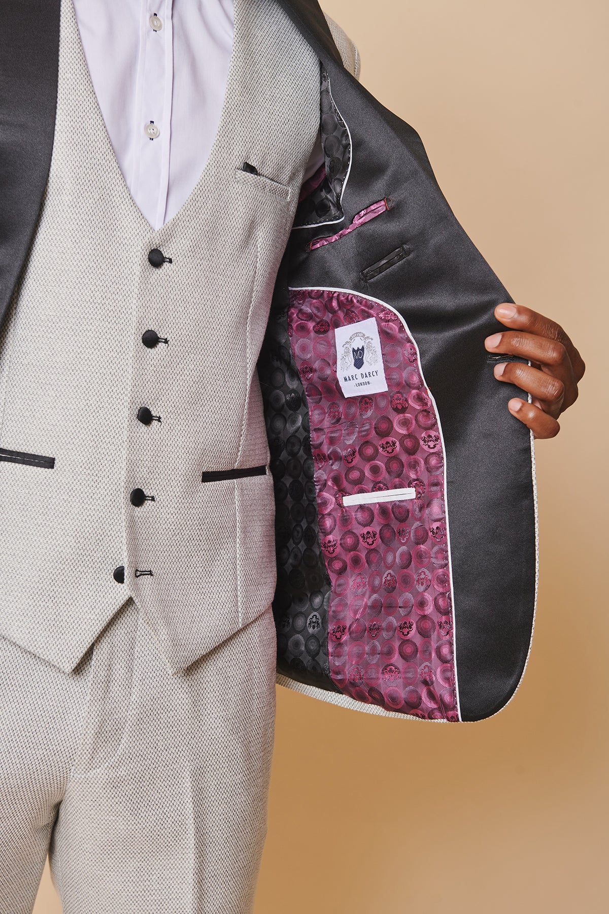 A man showcases the marcdarcy SPENCER - Stone Tux Lapel Blazer, revealing a satin purple skull-patterned lining with the DALEY QUARTZ label. The slim fit and beige backdrop add refined elegance for black tie events.
