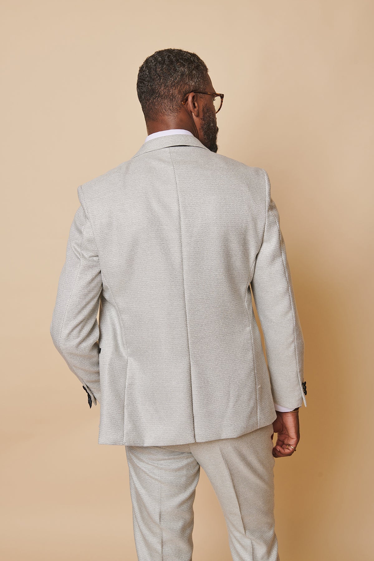 A man with short curly hair and glasses stands with his back to the camera in marcdarcy's The Everton Collection | Ashley Young In SPENCER Stone Tux Lapel Suit, set against a beige background, his left hand slightly bent at his side.
