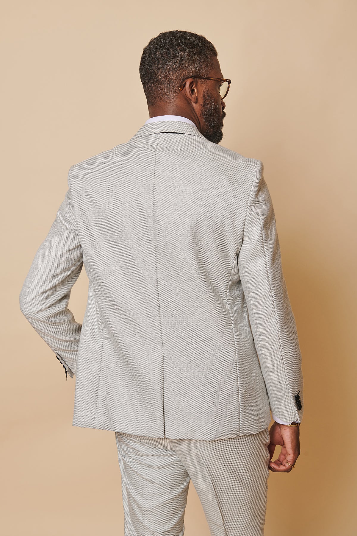 A man with short hair, beard, and glasses wears the marcdarcy SPENCER - Stone Tux Lapel Blazer in a slim fit, turned slightly to the side against a solid beige background—an ideal look for black tie events.