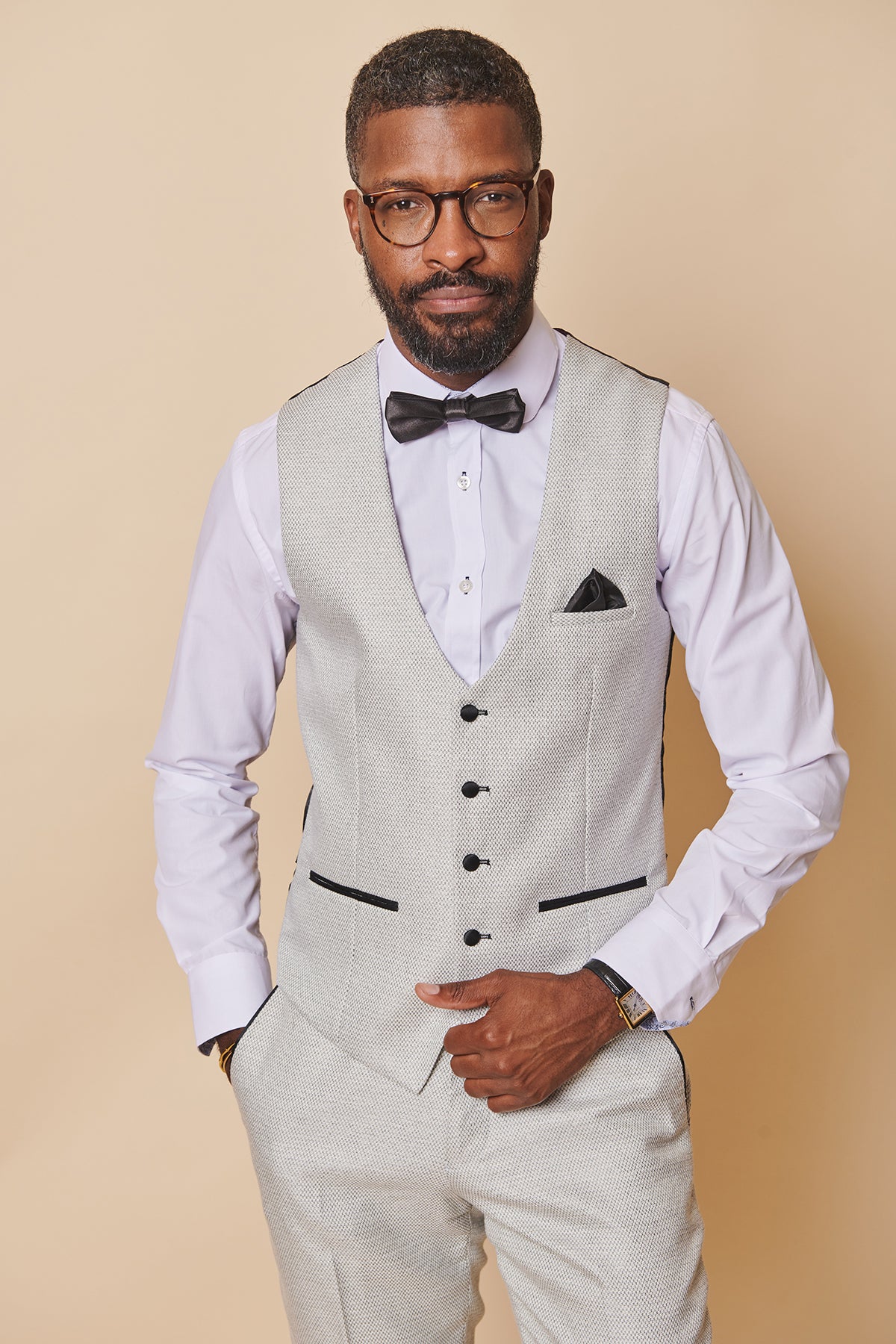 A man with short hair, a beard, and glasses wears the marcdarcy SPENCER Stone Single Breasted Waistcoat with matching pants, a white shirt, and black bow tie. He stands with hands in pockets against a beige background.