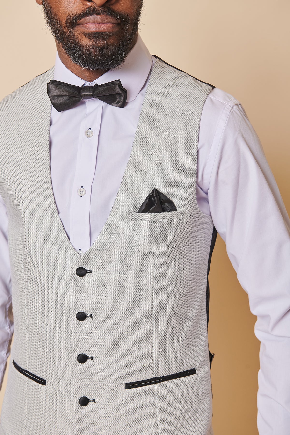 A man wears the marcdarcy SPENCER Stone Single Breasted Waistcoat with black buttons, paired with a white dress shirt, black bow tie, and black pocket square against a beige background. His face is partially visible.
