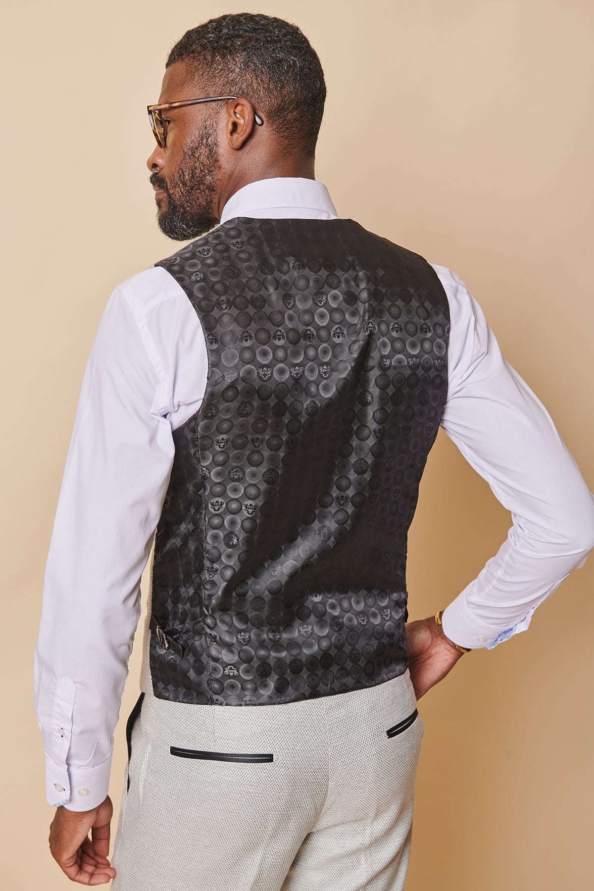 A man with short hair, a beard, and glasses stands with his back to the camera, wearing the marcdarcy SPENCER - Stone Single Breasted Waistcoat over a white shirt and light gray dress pants.