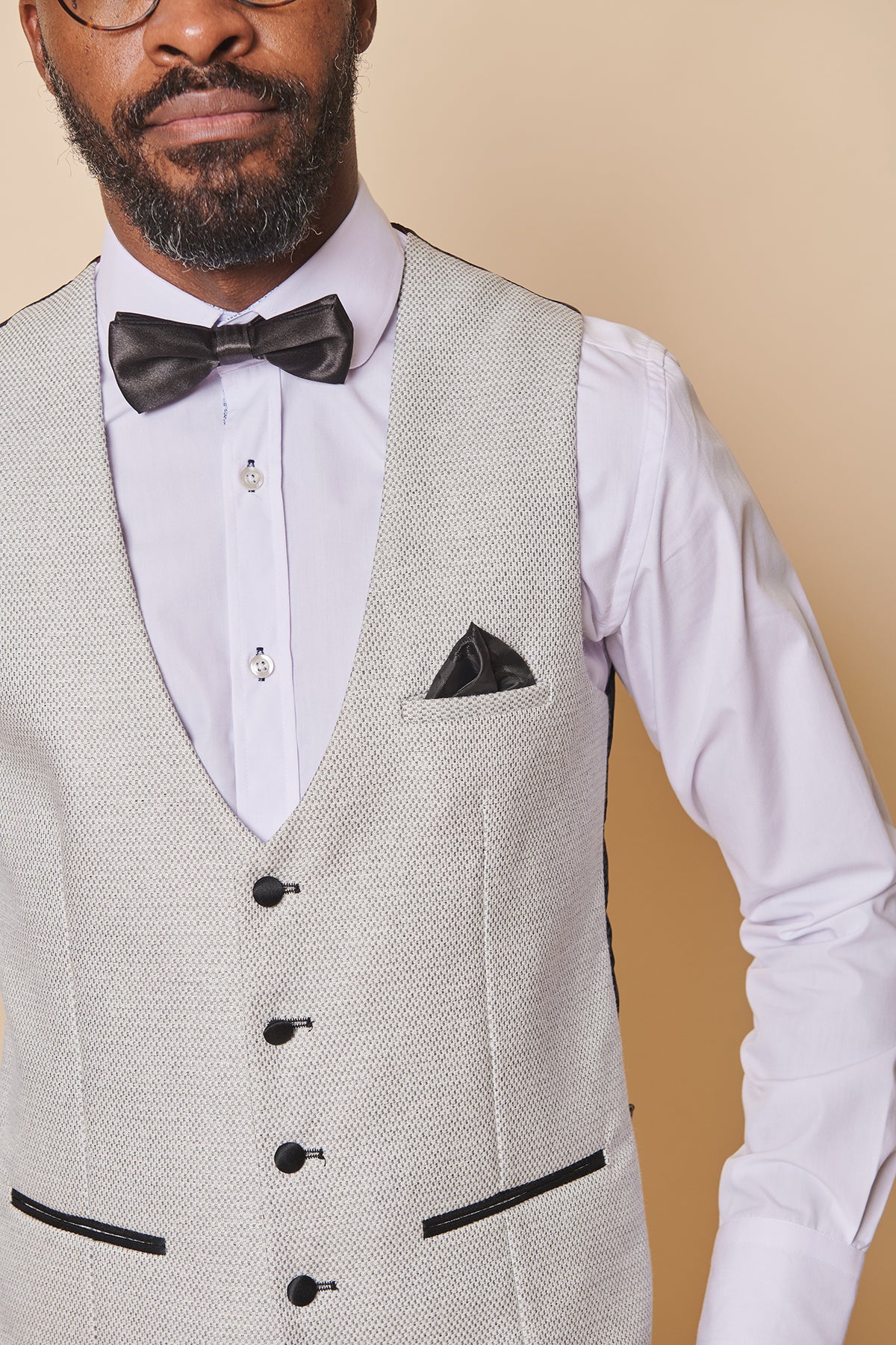 A man wearing the marcdarcy SPENCER Stone Single Breasted Waistcoat, with a white dress shirt, black bow tie, and matching pocket square, stands against a beige background. Only his lower face and upper body are visible.