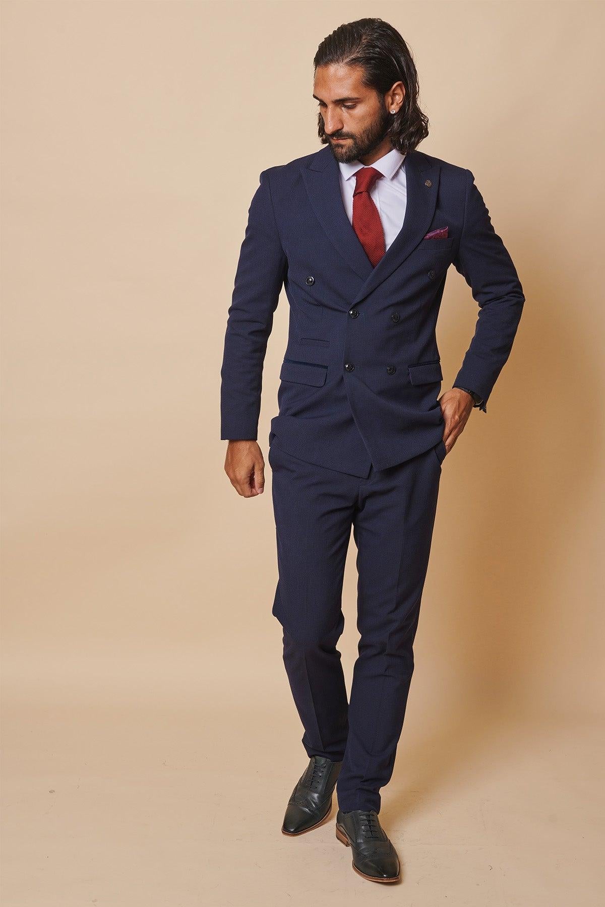 Former Sunderland footballer Michael Gray wears the Marc Darcy Bromley Navy Double-Breasted Suit, paired with a white shirt, red tie, and black shoes, standing against a beige background and looking down to his left.