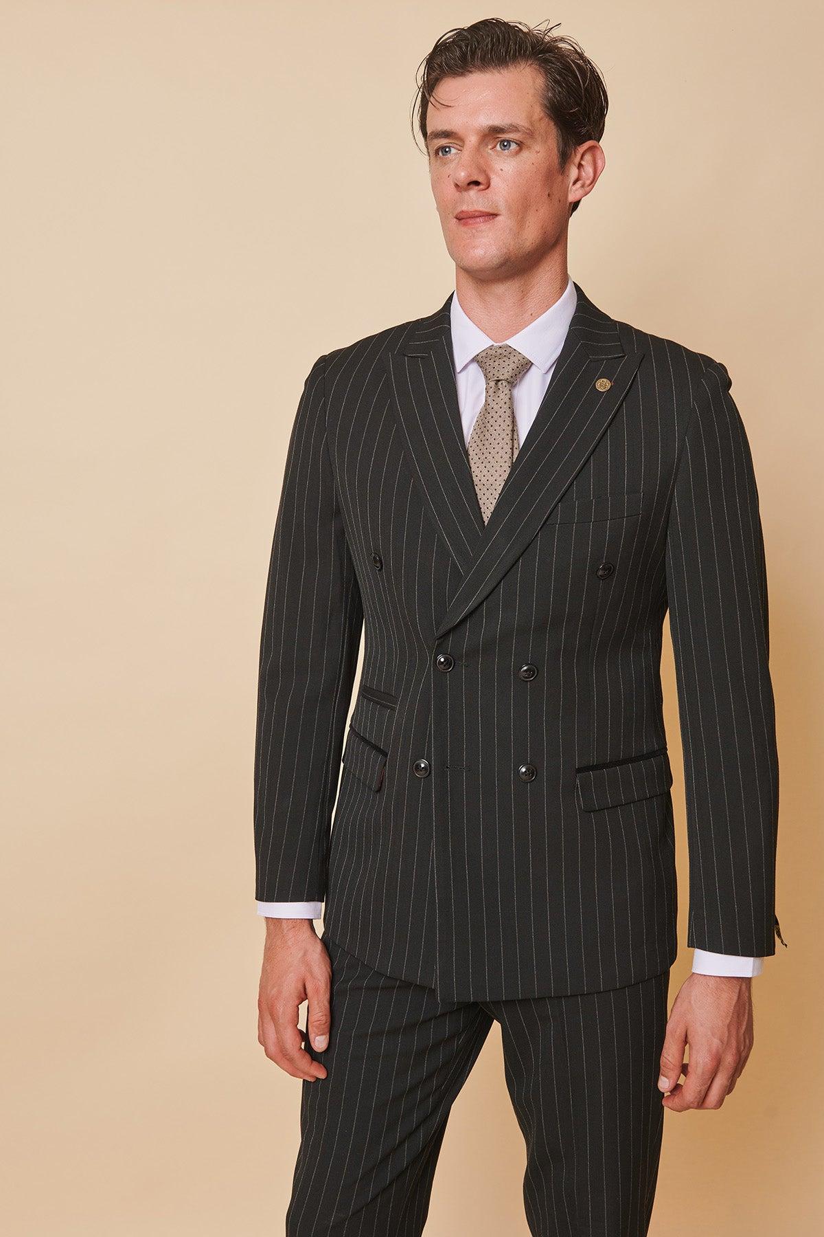 Coronation Street’s Mason Radcliffe (Luca Toolan) wears a Marc Darcy ROCCO Black Pinstripe Double Breasted Two Piece Suit, styled with a white shirt and patterned tie, standing against a beige background and looking composed.