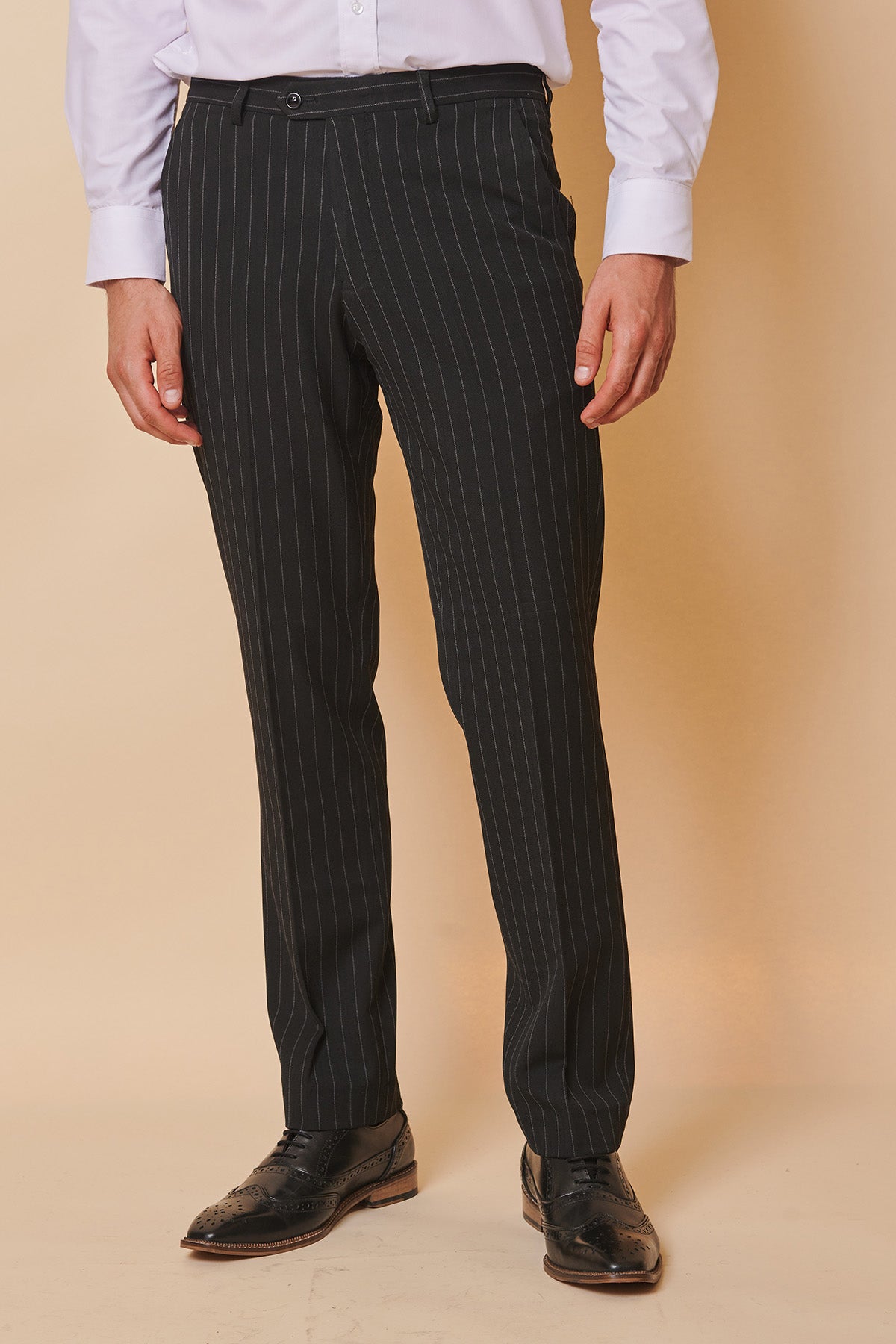 A man wears Marc Darcy's ROCCO Black Pinstripe Trousers and a white dress shirt, standing against a cream background. His look is finished with black wingtip shoes.