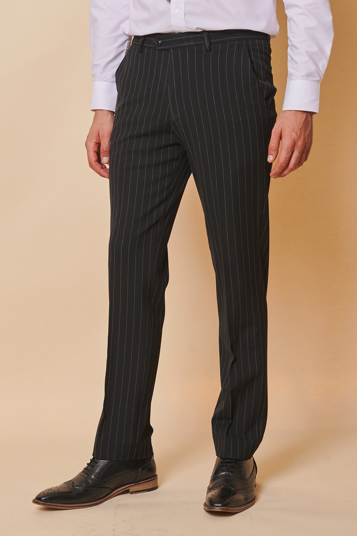 A person models the Marc Darcy ROCCO Black Pinstripe Trousers against a beige background, paired with a white dress shirt and black dress shoes. Their face is not visible.