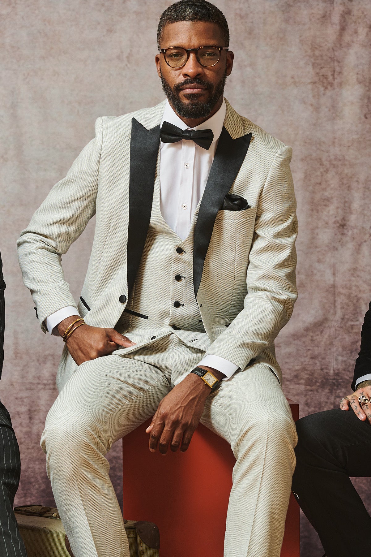 A bearded man in glasses sits on a red block, wearing the marcdarcy SPENCER Stone Tux Lapel Three Piece Suit with a white shirt, black bow tie, and watch, set against a textured studio backdrop.