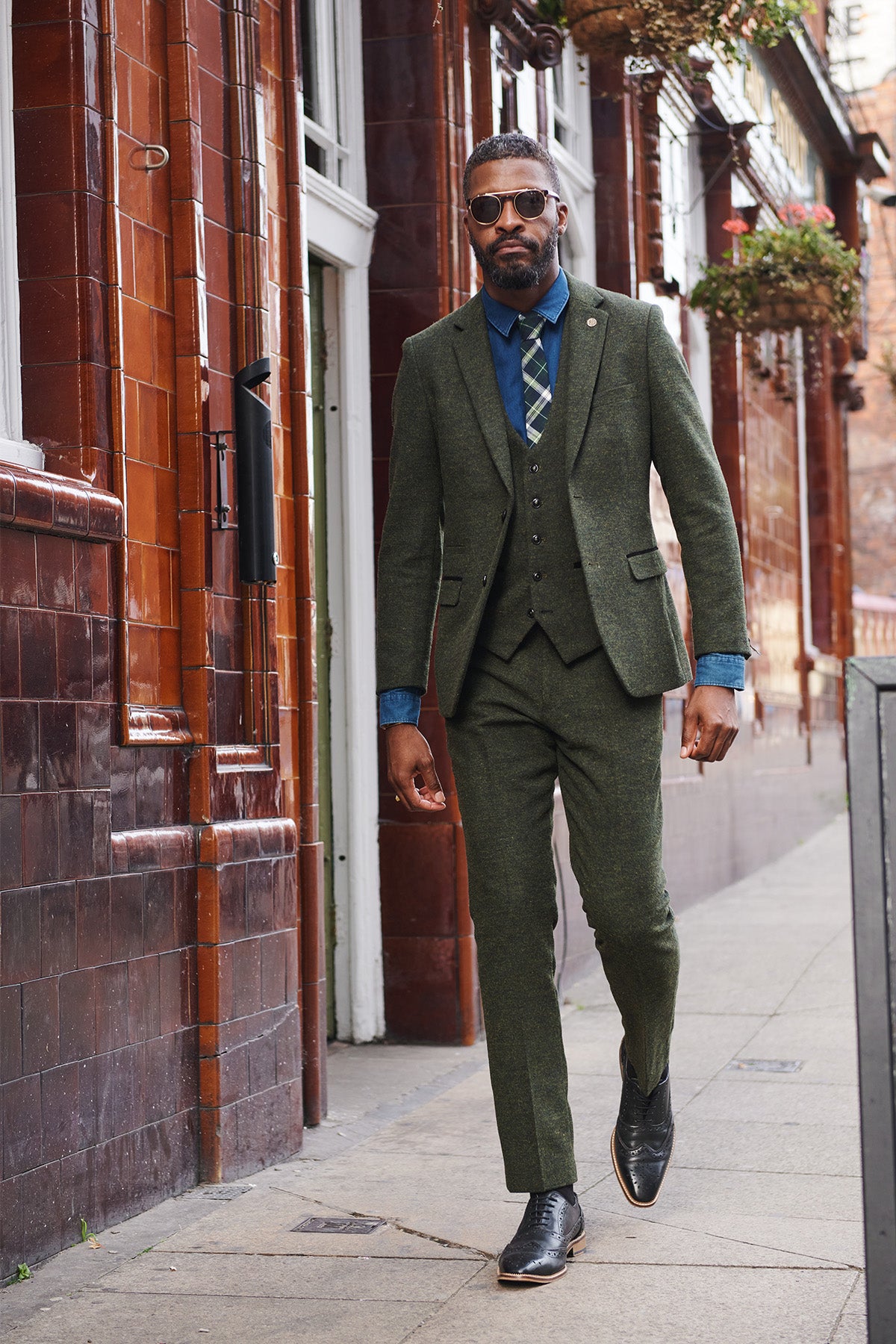 A man in a marcdarcy MARLOW Olive Green Tweed Three Piece Suit, blue shirt, plaid tie, and sunglasses strides confidently along a city sidewalk next to a red brick building with hanging plants.