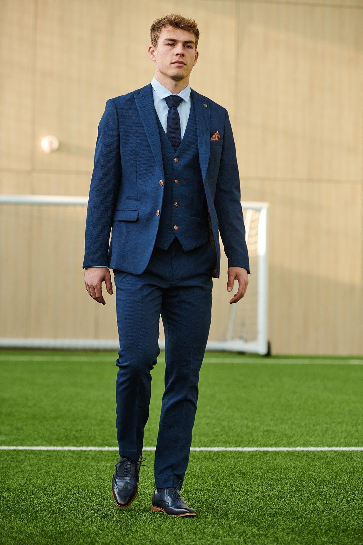 A young man in the marcdarcy MAX Royal Blue Suit from The Leicester City F.C. Collection, as worn by Mads Hermansen, walks confidently on a green sports field with a goalpost and tan wall in the background.
