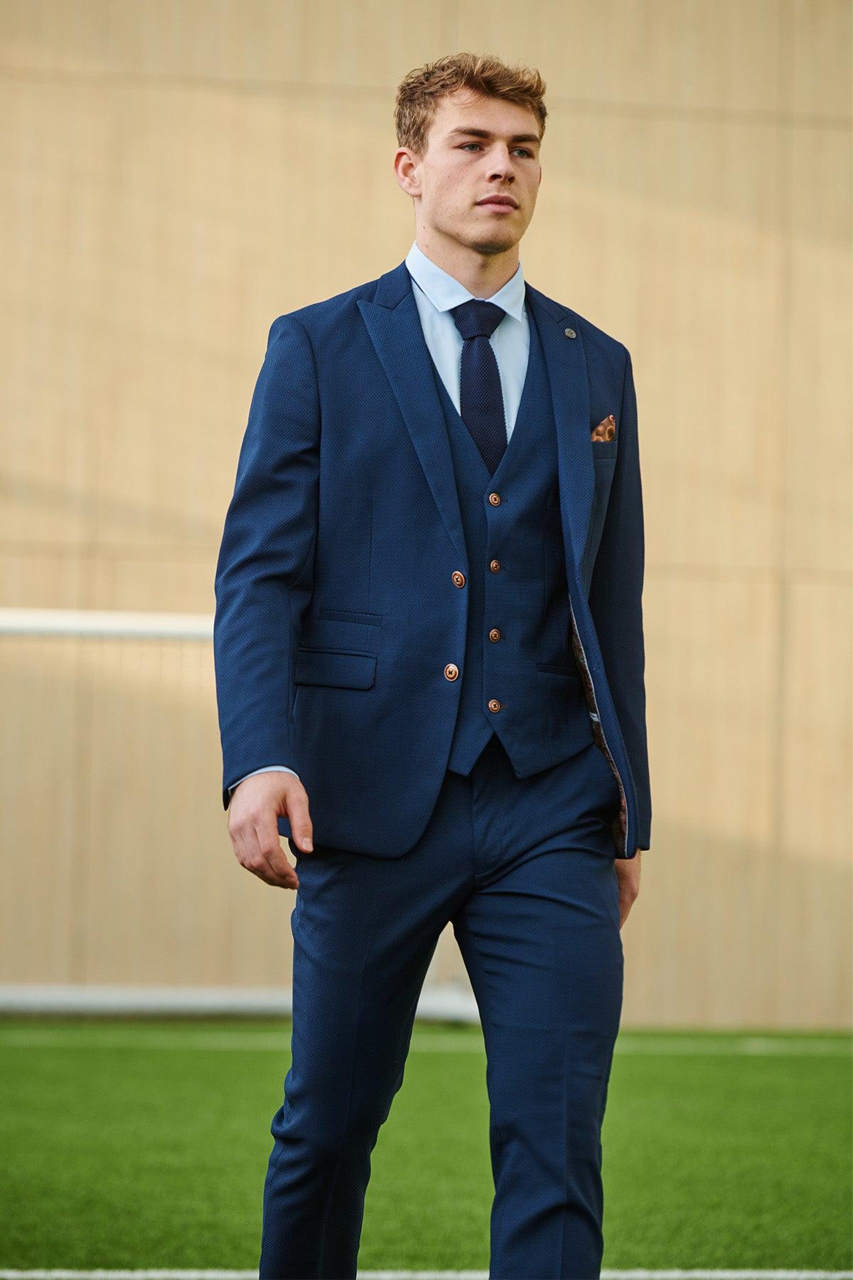 A young man confidently walks outdoors on green artificial grass wearing the marcdarcy Leicester City F.C. Collection - MAX Royal Blue Suit as worn by Mads Hermansen, with a beige building in the background.
