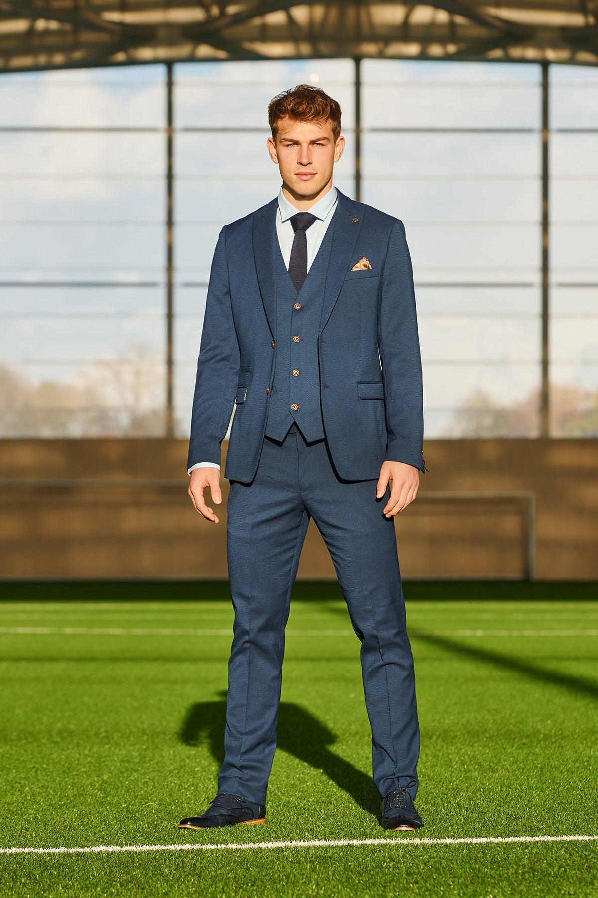 A man wearing the marcdarcy Official Leicester City F.C. Club Suit | MAX Royal Blue Suit stands confidently on a green indoor soccer field, sunlight streaming through large windows in the background.