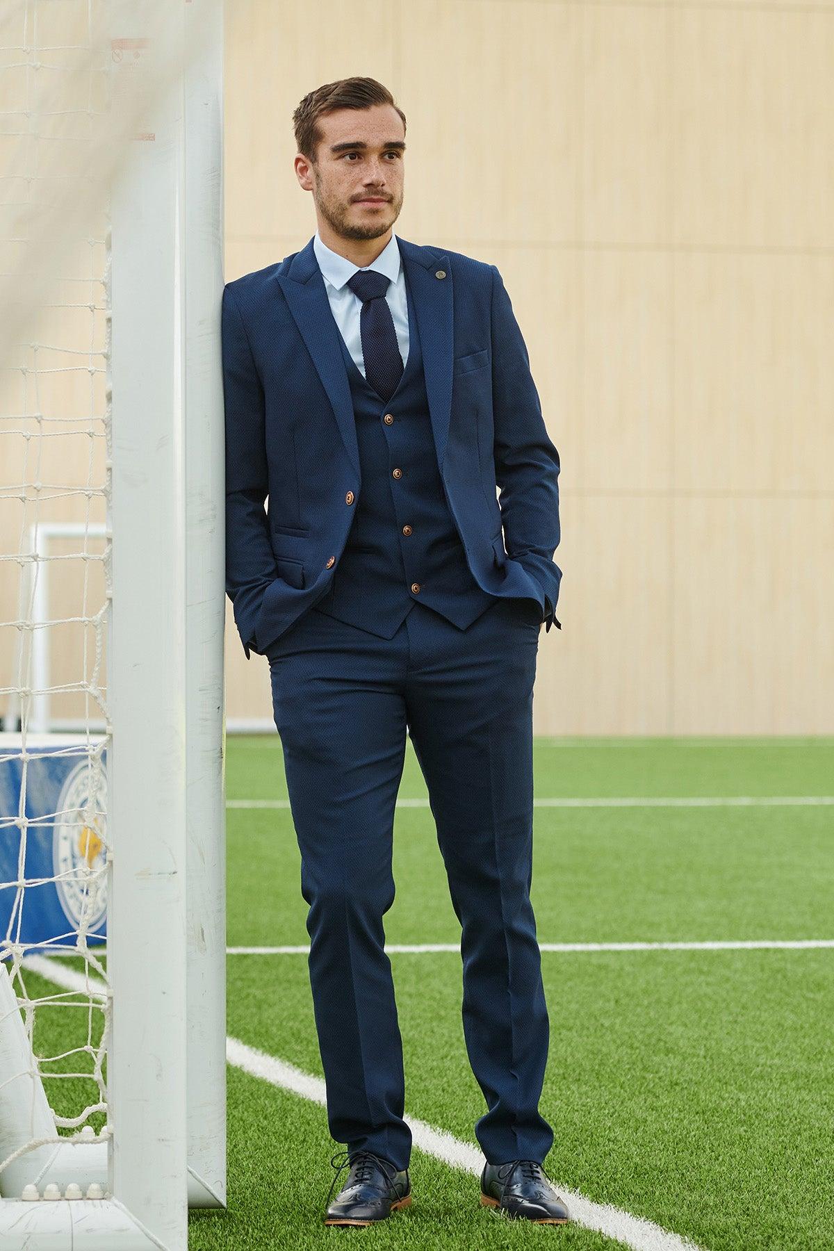 A man wearing the marcdarcy Official Leicester City F.C. Club Suit | MAX Royal Blue Suit stands with hands in pockets, leaning against a soccer goalpost on a turf field.