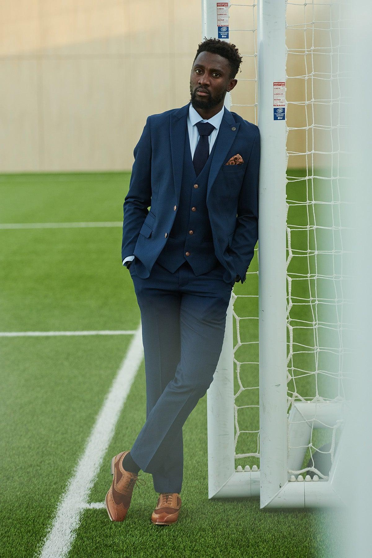 A man in the marcdarcy Leicester City F.C. Collection - MAX Royal Blue Suit, as worn by Wilfred Ndidi, and brown shoes leans against an outdoor soccer goalpost, looking relaxed with his hands in his pockets.