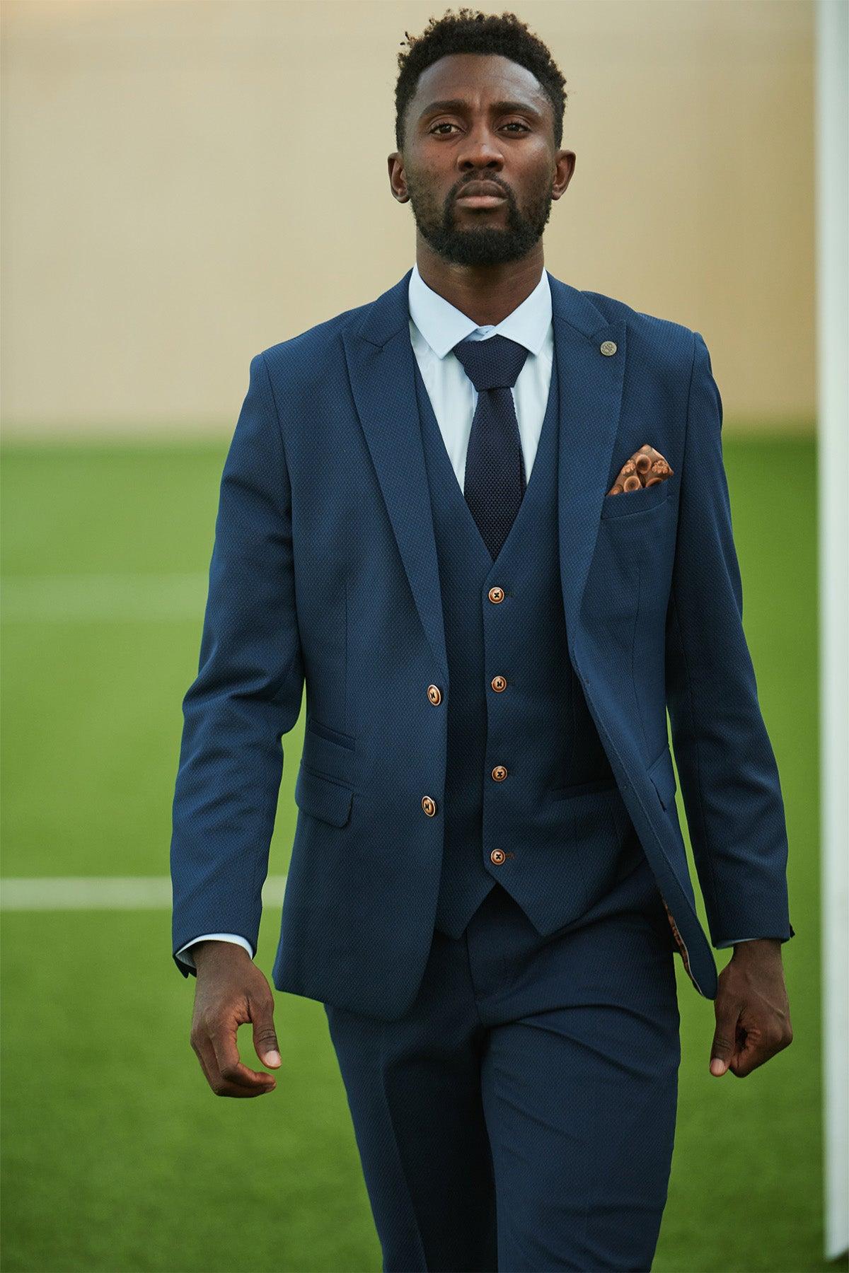 A man wearing the marcdarcy Leicester City F.C. Collection - MAX Royal Blue Suit, as worn by Wilfred Ndidi, walks confidently on a football field. His slim fit blazer and composed look highlight his formal style against the blurred background.