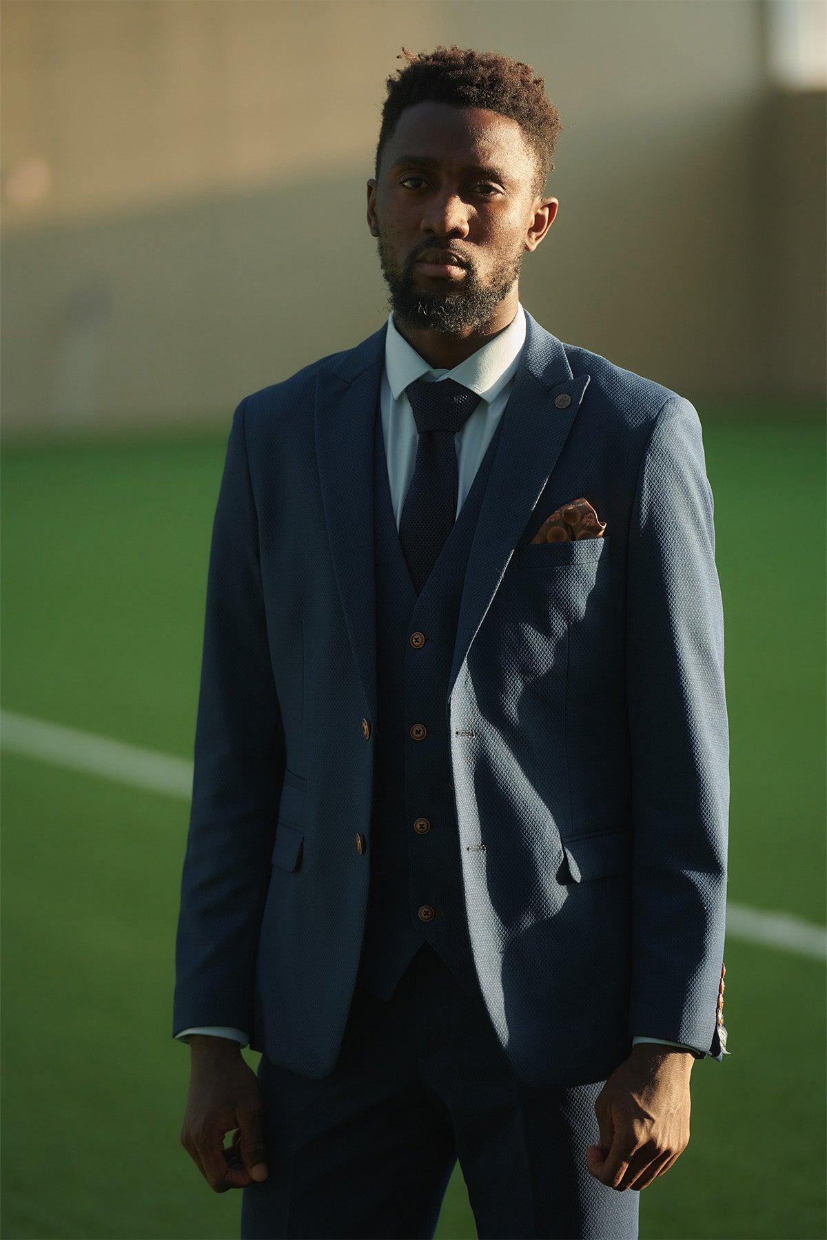 A man wears the marcdarcy Leicester City F.C. Collection MAX Royal Blue Suit as worn by Wilfred Ndidi—three-piece with white shirt and navy tie—standing on a sunny grass field, looking at the camera seriously.