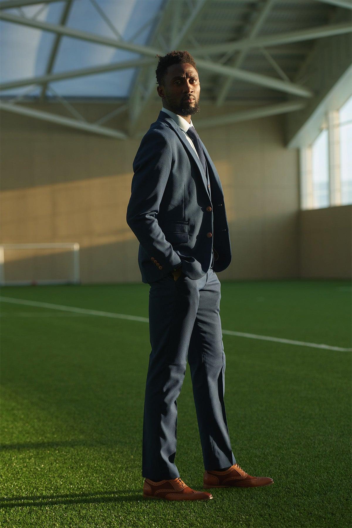 A man wears the marcdarcy Leicester City F.C. Collection MAX Royal Blue Suit as worn by Wilfred Ndidi, with tan shoes, standing hands-in-pockets on green turf.