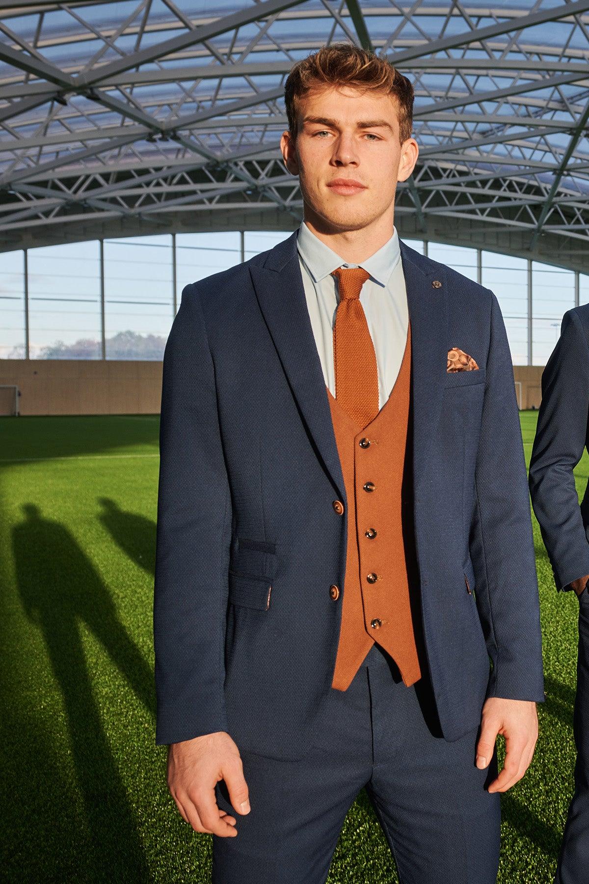 A young man stands on an indoor sports field, wearing the marcdarcy Official Leicester City F.C. Club Suit—MAX Royal Blue Suit with Kelvin Tan Waistcoat—as sunlight casts long shadows over the green artificial turf behind him.