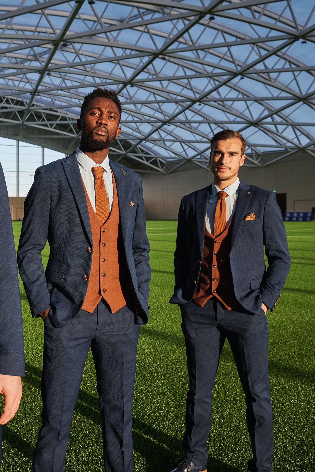 Two men stand on artificial turf in a large indoor sports facility, wearing the marcdarcy Official Leicester City F.C. Club Suit—MAX Royal Blue Suit paired with Kelvin Tan Waistcoat—sunlight streaming through the glass roof illuminates their stylish look.