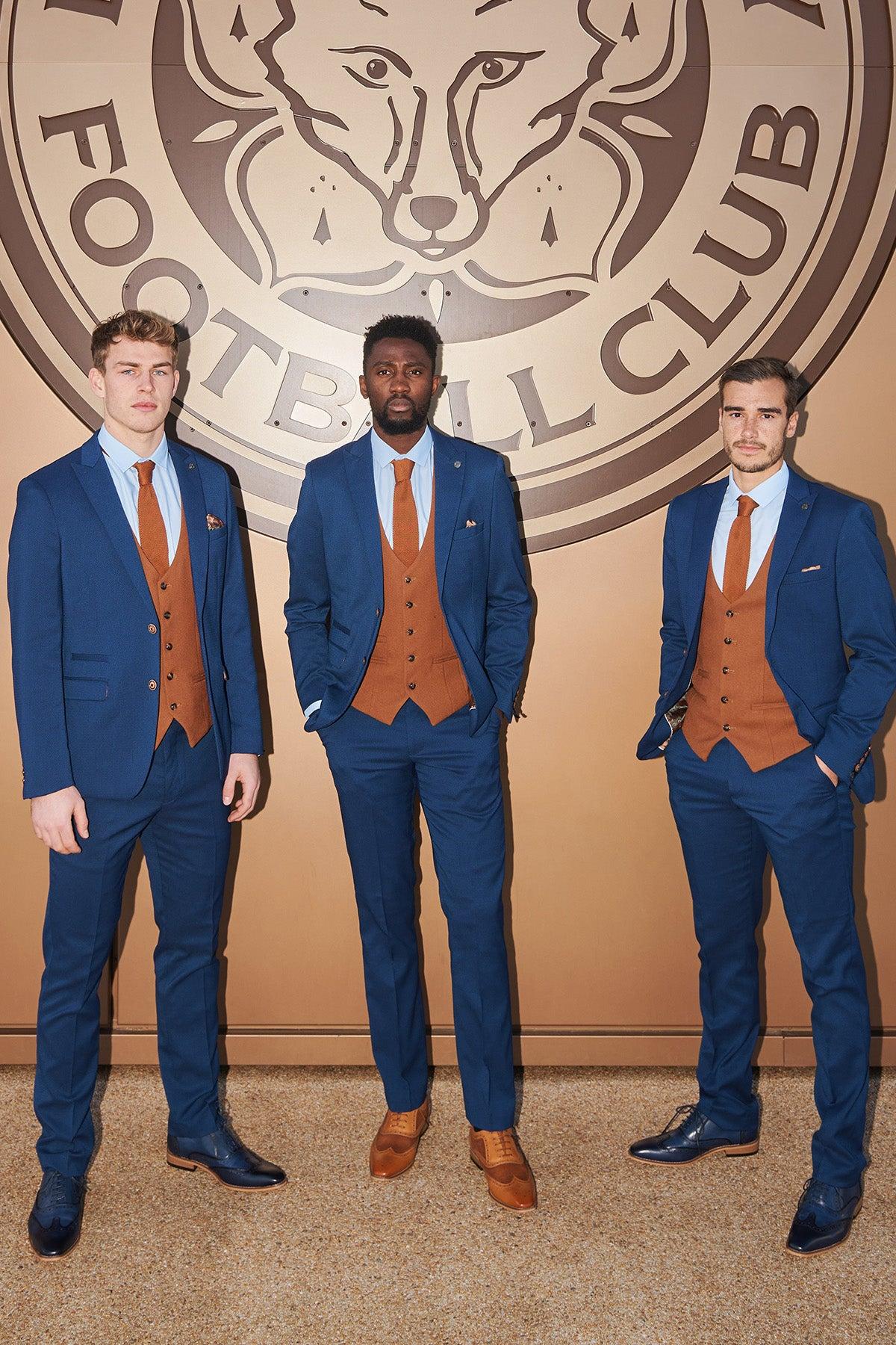 Three men wear marcdarcy Official Leicester City F.C. Club Suits—MAX Royal Blue Suits with Kelvin Tan Waistcoats—standing confidently, hands in pockets, before a large wolf-head football emblem on a tan background.
