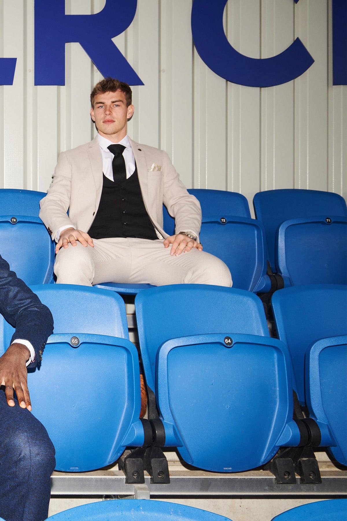 A man in the Marc Darcy Leicester City F.C. Collection HM5 Stone Suit with Kelvin Black Waistcoat, as worn by Mads Hermansen, sits among blue stadium seats, wearing a white shirt, black tie, and pocket square.