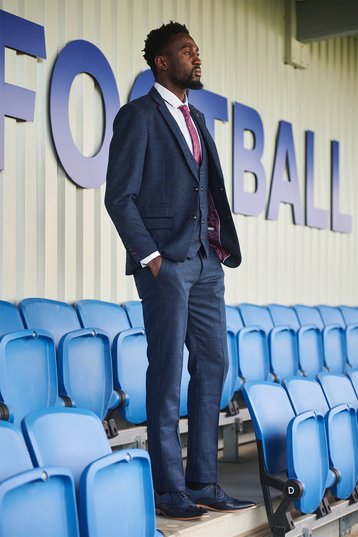 A man stands among blue stadium seats wearing the marcdarcy Leicester City F.C. MARLOW Navy Tweed Suit as worn by Wilfred Ndidi, with a white shirt, pink tie, and a FOOTBALL sign behind him.