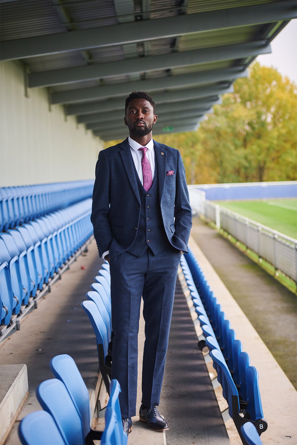 A man in a MARLOW Navy Tweed Suit from the Leicester City F.C. Collection by marcdarcy stands with hands in pockets on a stadium walkway, blue seats to his left and a green field to his right, wearing a pink tie.