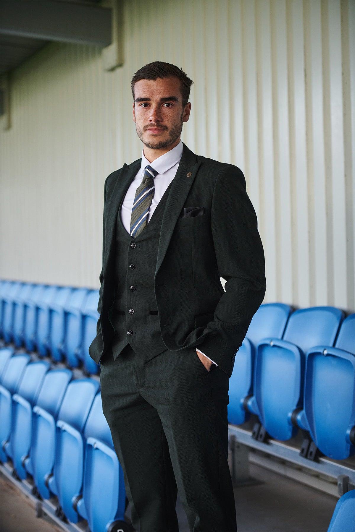 A man in the Marc Darcy Leicester City F.C. Collection BROMLEY Olive Green Check Suit, as worn by Harry Winks, stands with one hand in his pocket beside empty blue stadium seats and a corrugated metal wall.